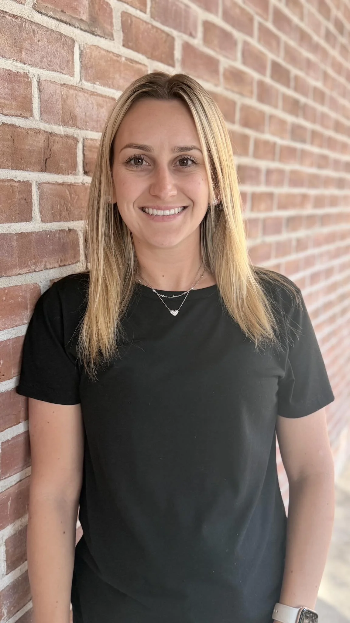 A woman with blonde hair, smiling, wearing a black t-shirt, standing against a red brick wall.
