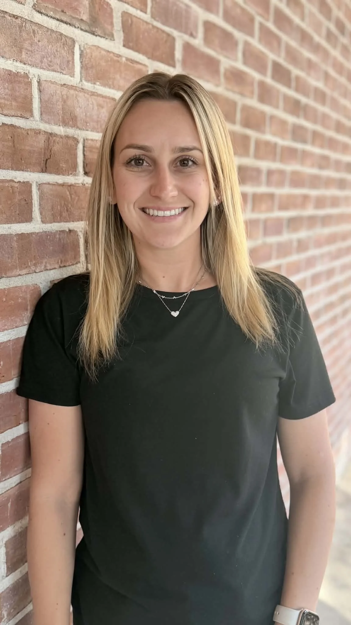 A smiling woman with blonde hair wearing a black t-shirt and necklaces, standing next to a brick wall.