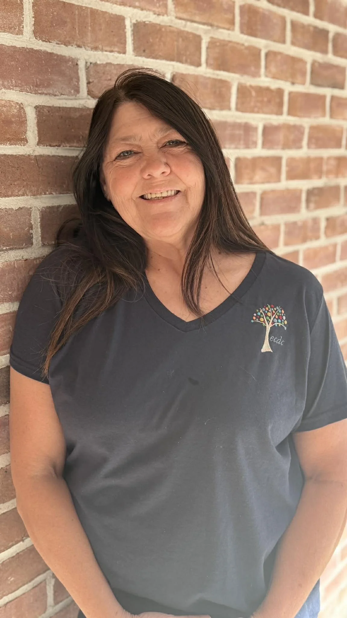 A woman with dark brown hair, wearing a black T-shirt with a colorful tree logo, leaning against a brick wall and smiling.