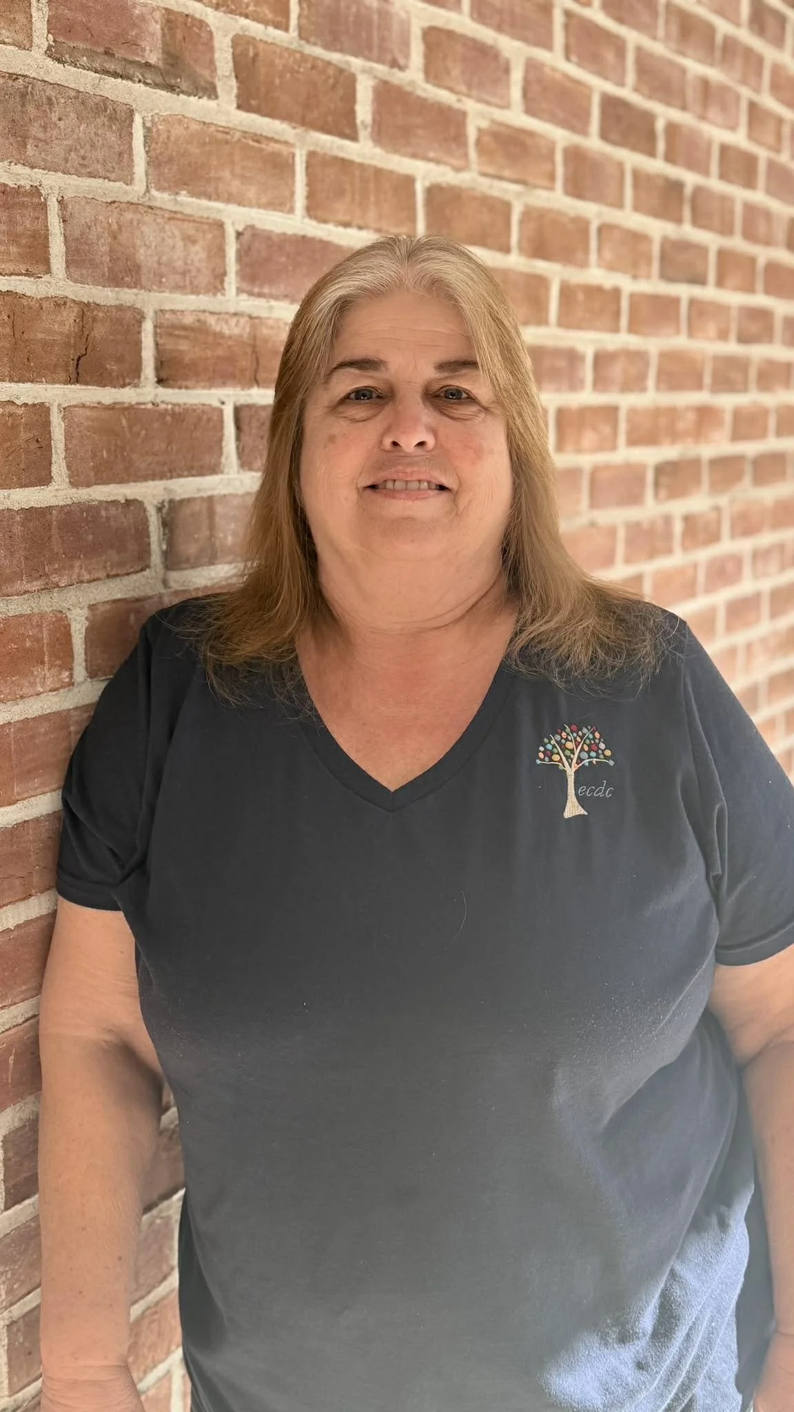 A woman with shoulder-length brown hair smiling, standing against a red brick wall, wearing a black V-neck T-shirt with a colorful embroidered tree and the initials "e.c." on the upper left side.