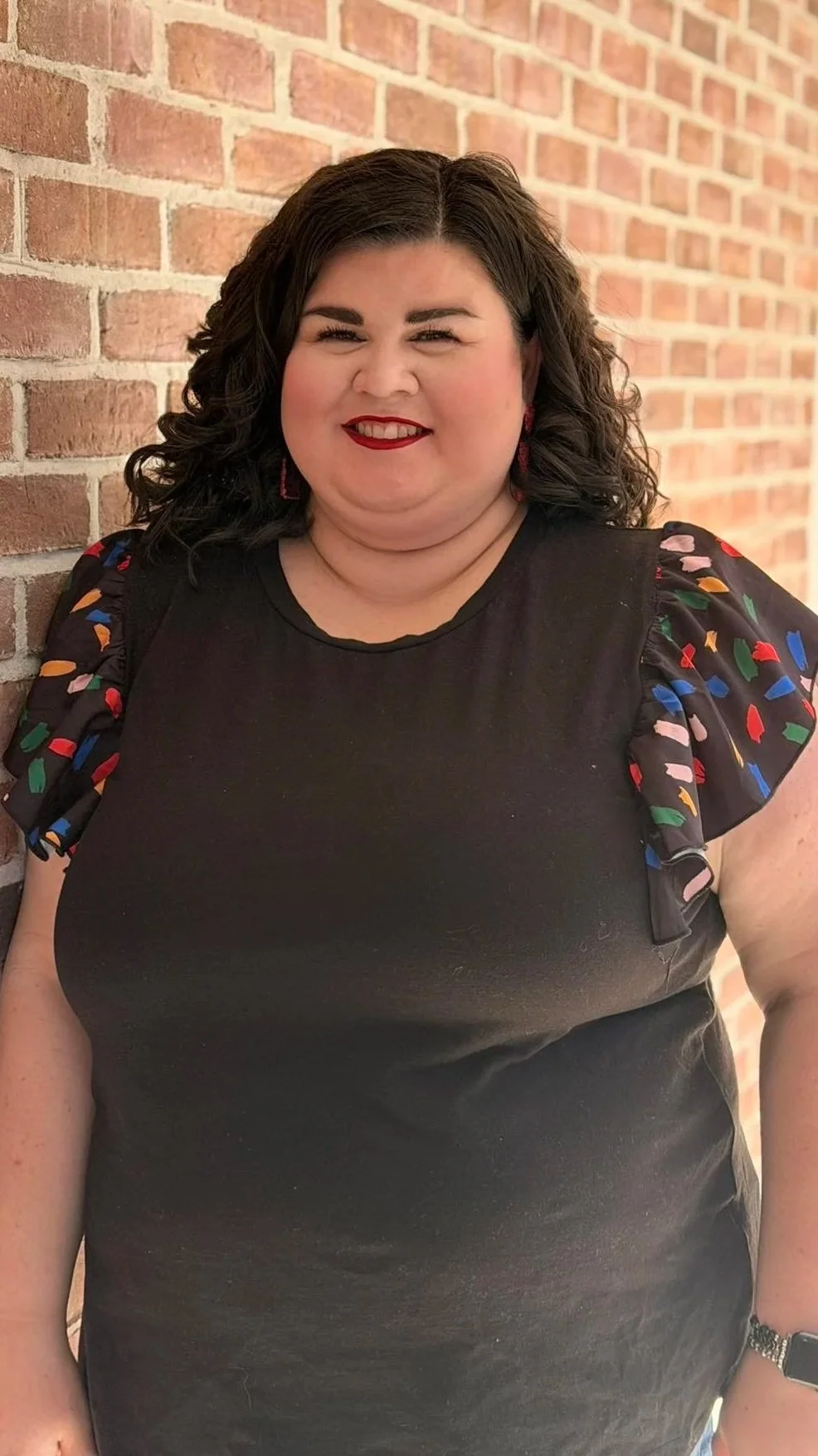 A woman with dark curly hair and red lipstick leaning against a brick wall, wearing a black top with colorful puffed sleeves.