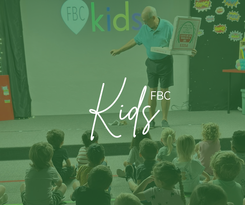 An elderly man in a teal polo and dark shorts standing on stage in front of a group of children, holding an open pizza box. The stage has a colorful backdrop with the words 'FBC kids' and various comic-style graphics. The children are sitting on the floor, listening to the man. A table with colorful items is on the right side.
