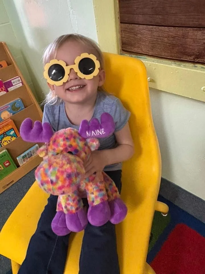 A young girl sitting on a yellow chair, wearing large yellow sunglasses with black lenses, and purple mittens with the word 'MAINE' on them. She is holding a colorful plush reindeer toy with purple feet and antlers, and smiling.