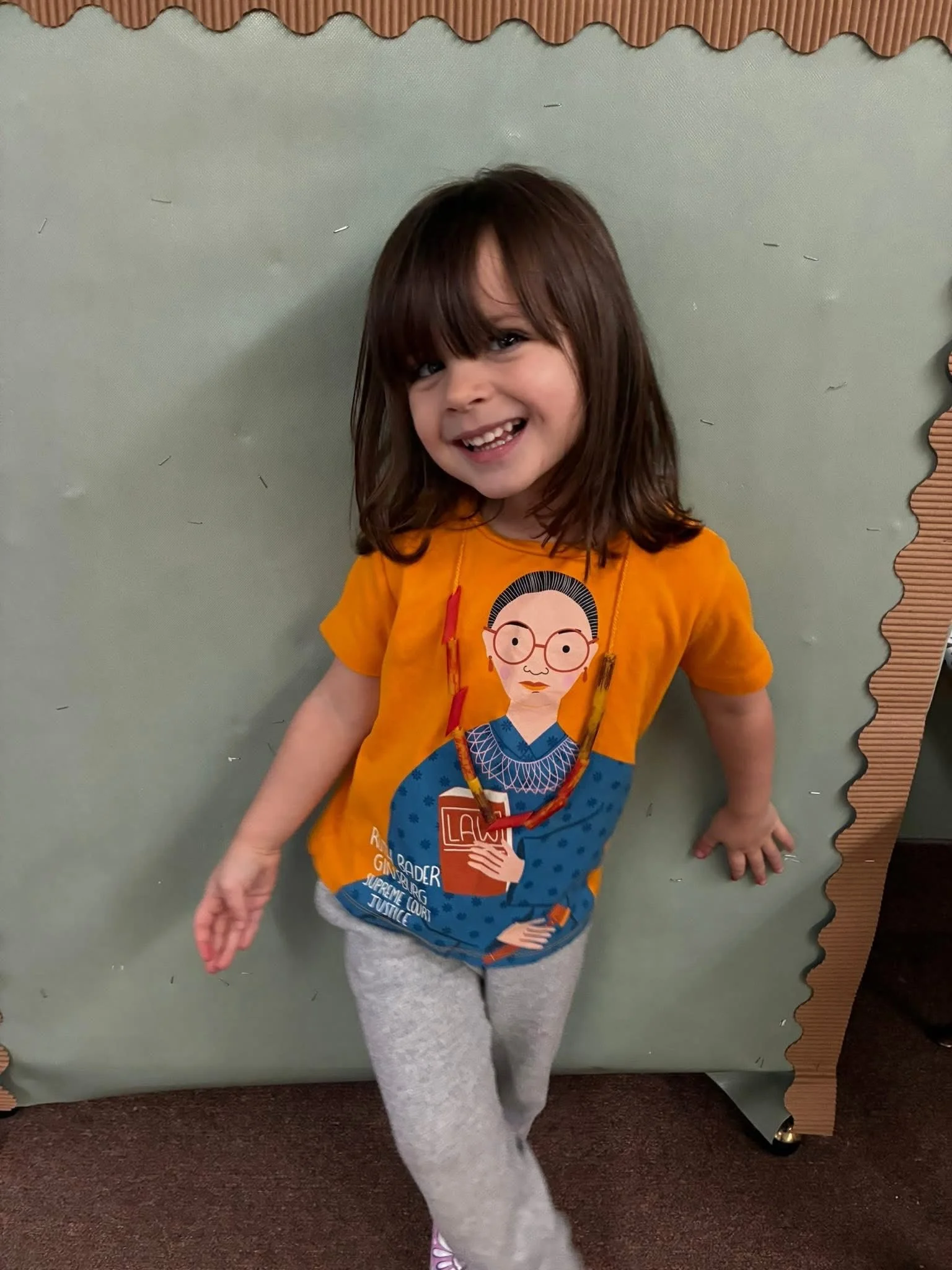 A young girl with brown hair and a big smile, standing against a green wall with a scalloped border, wearing a graphic orange T-shirt with a cartoon woman holding a law book, gray pants, and pink shoes.
