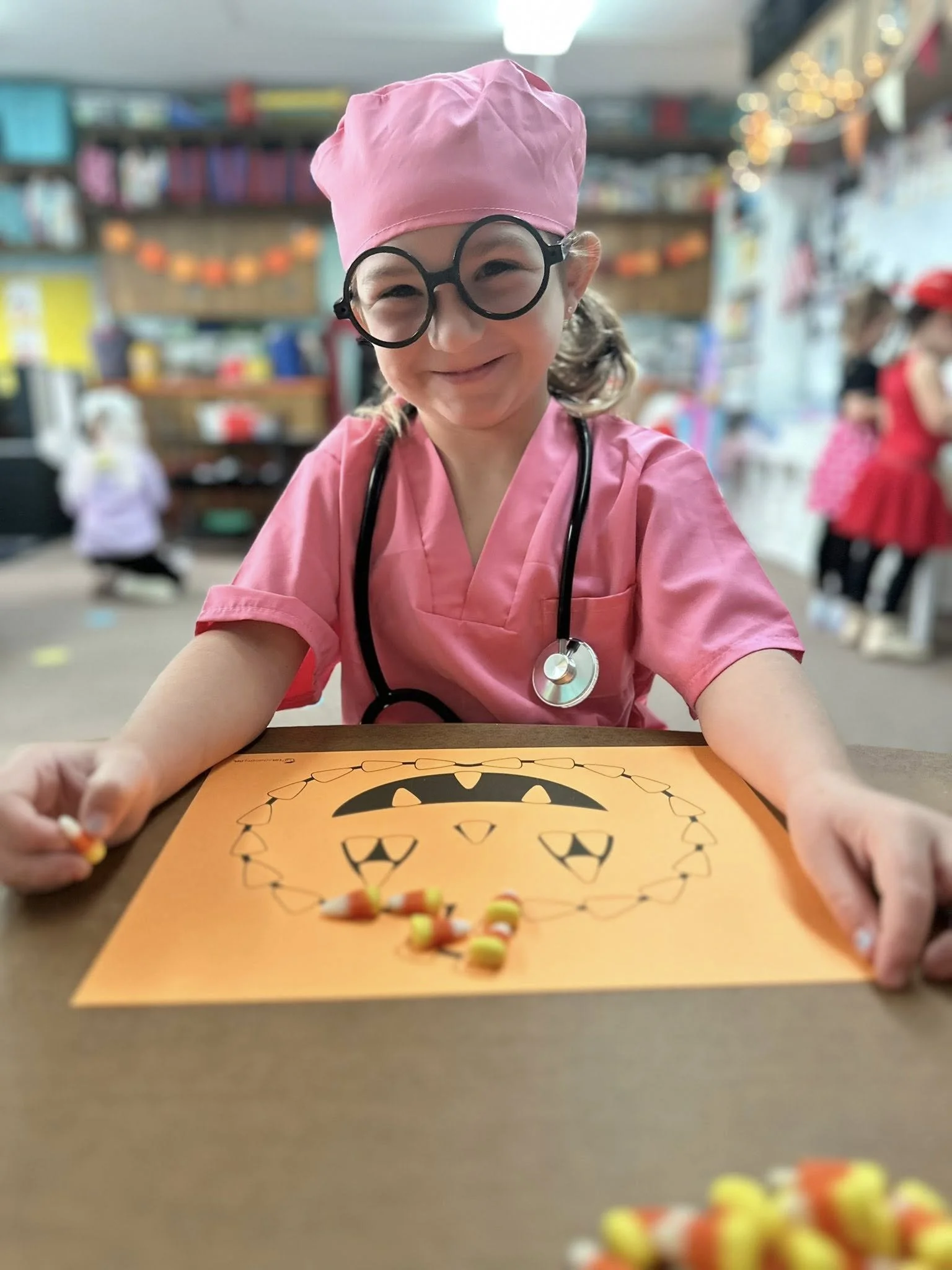A young girl dressed as a doctor, wearing pink scrubs, a pink cap, and oversized glasses, smiling at the camera while sitting at a table with a piece of orange paper displaying a Halloween-themed drawing of a smiling pumpkin face, in a classroom or party setting.