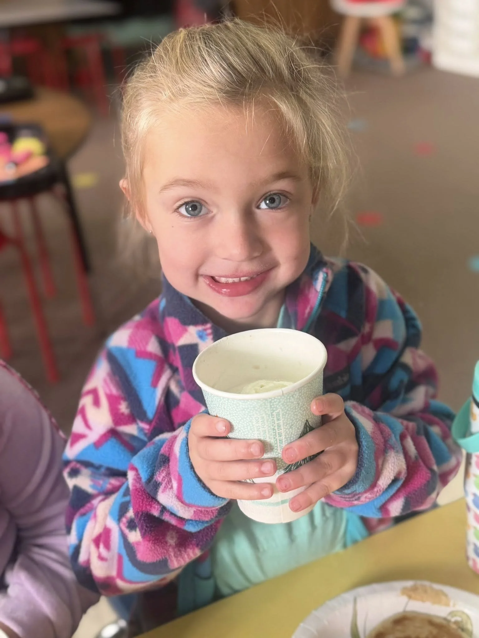 A young girl with blonde hair and blue eyes holding a cup of ice cream, smiling at the camera.