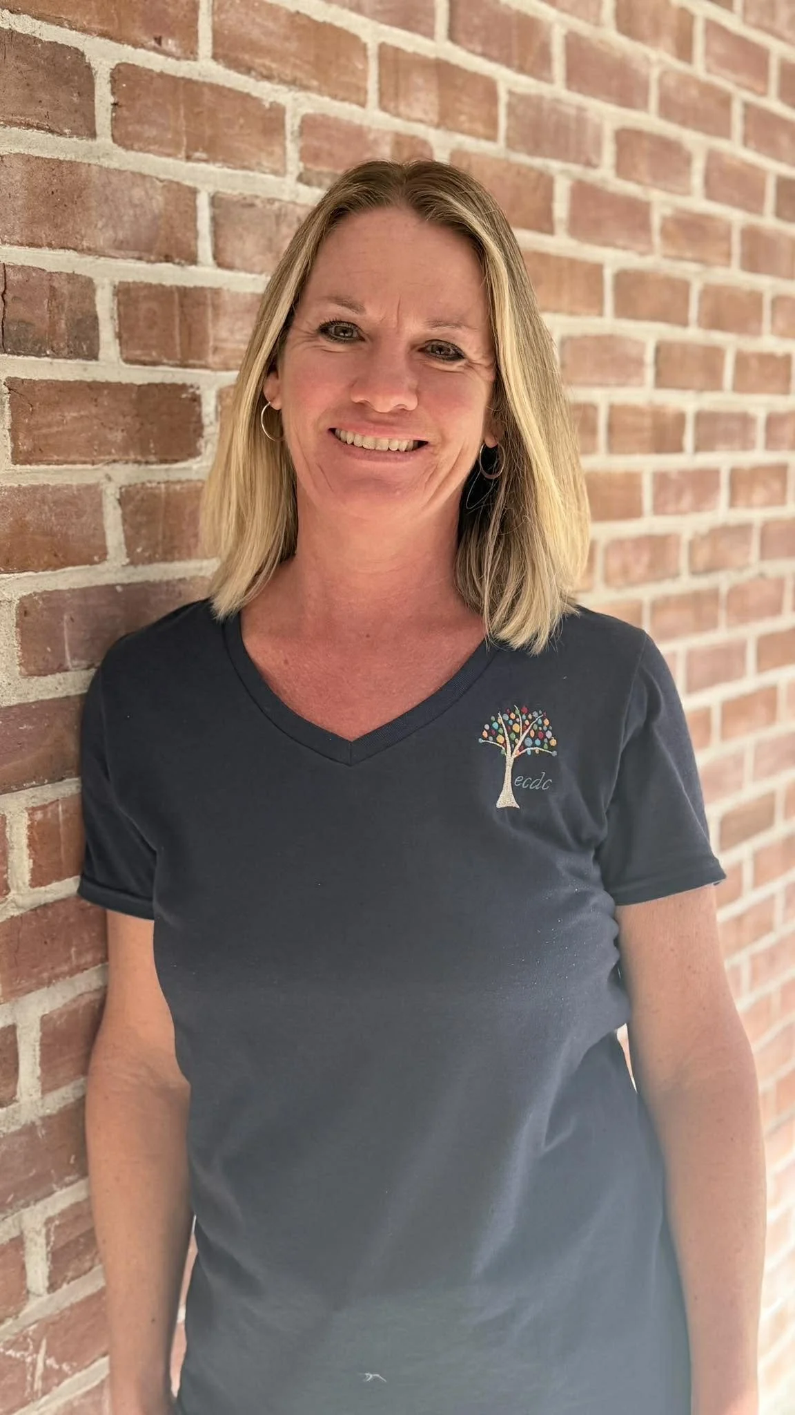 A smiling woman with blonde hair, wearing earrings and a navy blue t-shirt with a colorful tree logo, standing against a brick wall.