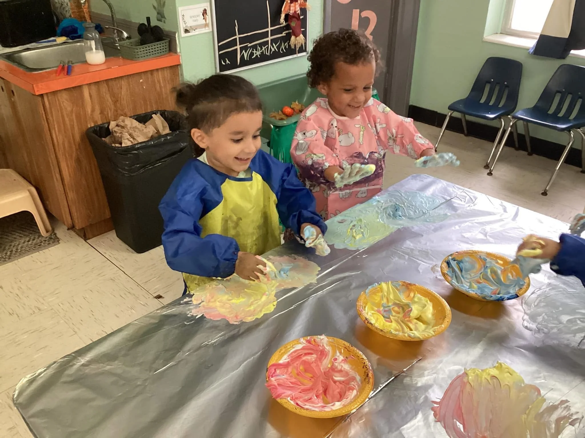 Two children smiling and playing with colorful paint on a large sheet of paper in a classroom, with bowls of paint on the table.
