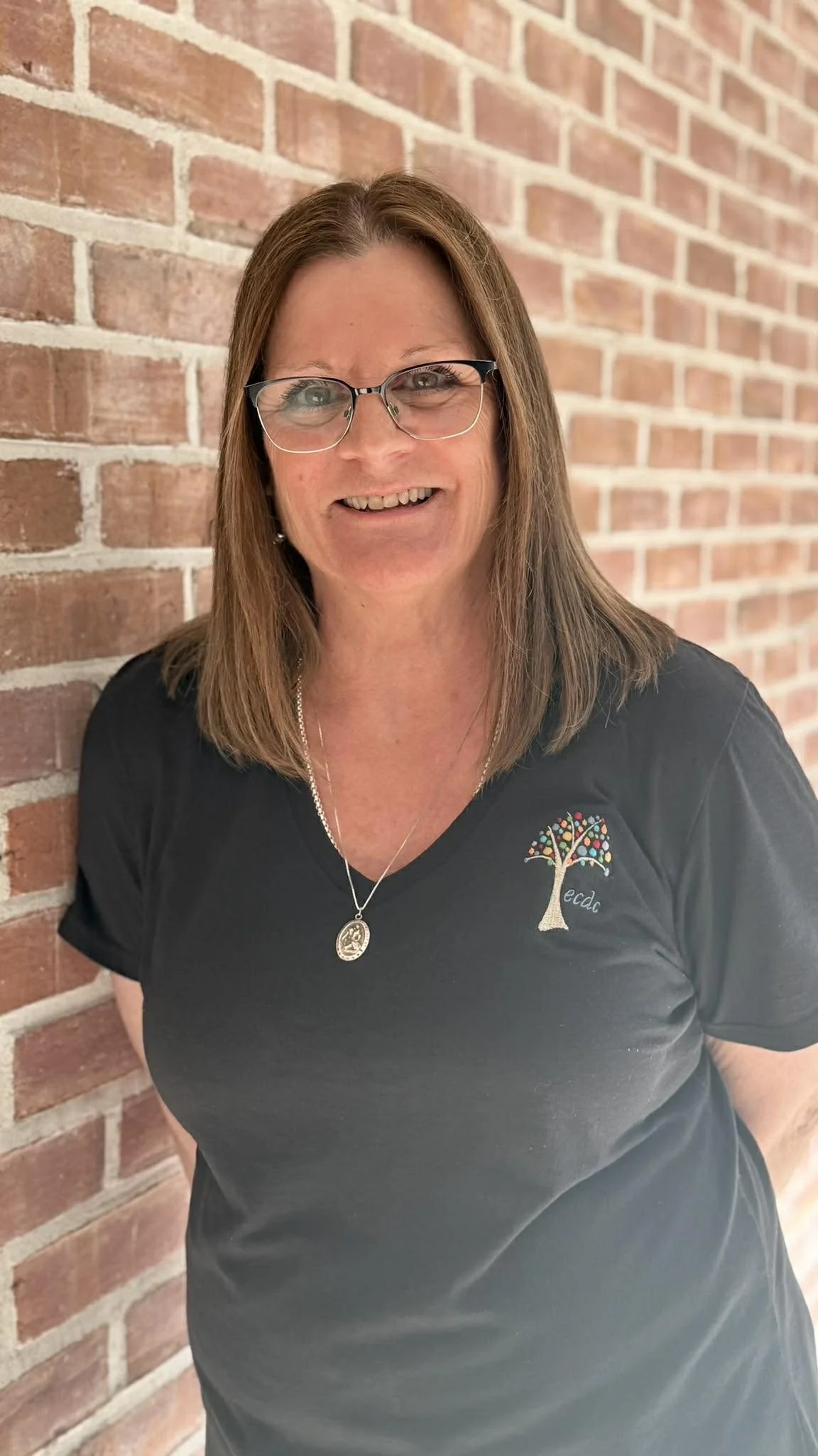 A smiling woman with glasses and brown hair, wearing a black t-shirt with a colorful tree design and the word 'ecda' on it, standing in front of a brick wall.