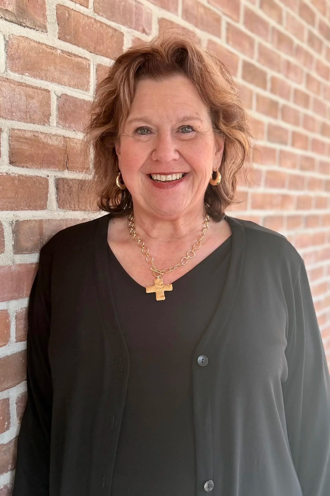 Smiling woman with shoulder-length reddish-brown hair, wearing gold jewelry including hoop earrings and a chain necklace with a cross pendant, dressed in black, standing against a brick wall.