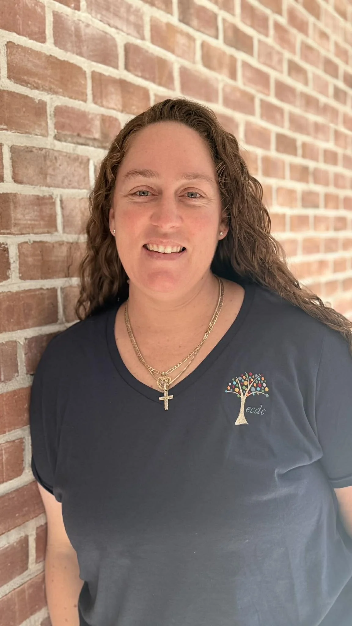 A woman with long curly hair smiling, standing against a brick wall, wearing a dark shirt with a colorful tree logo and gold jewelry.