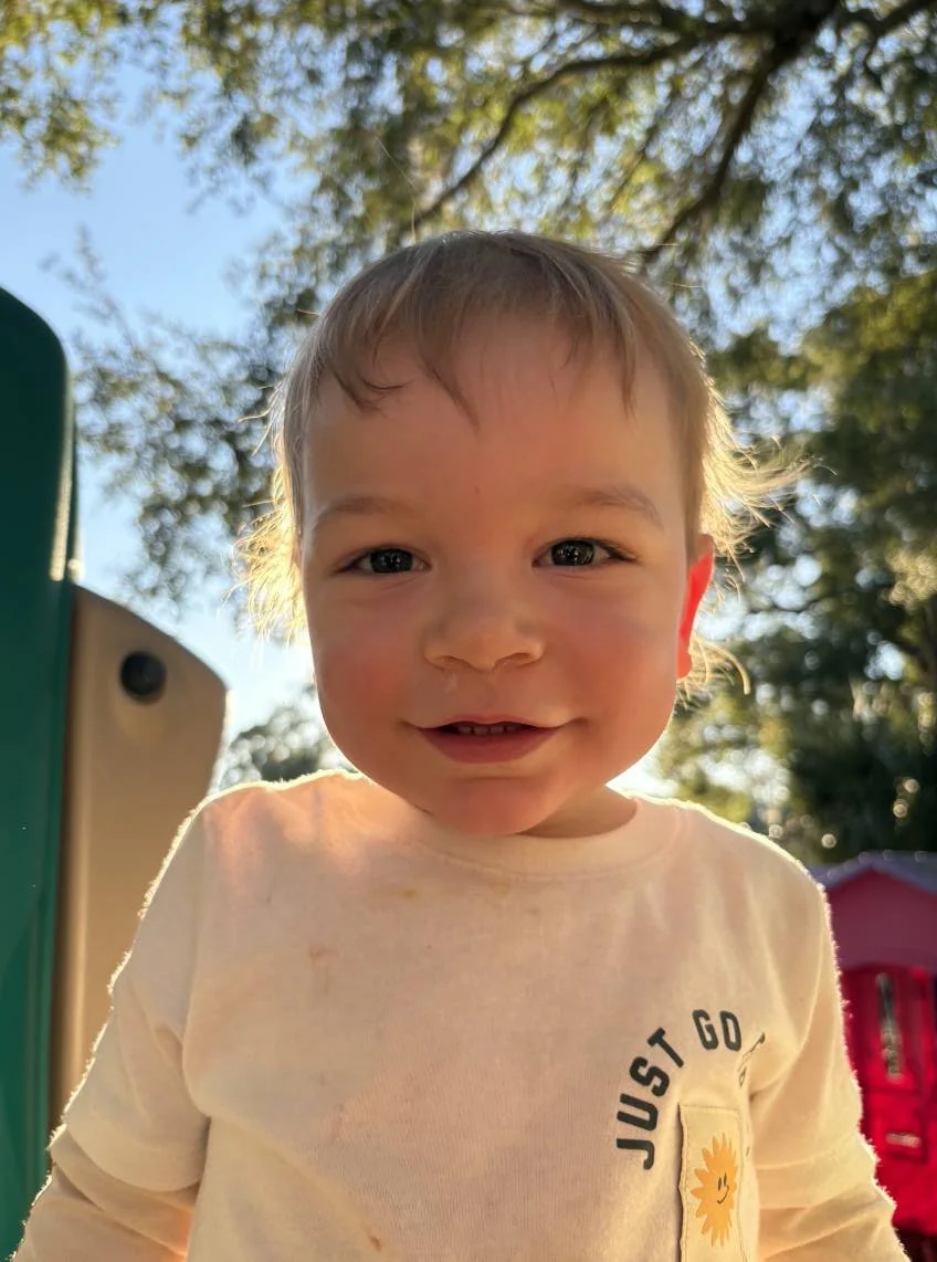 A young boy with light brown hair smiling outdoors with trees and a clear sky in the background.