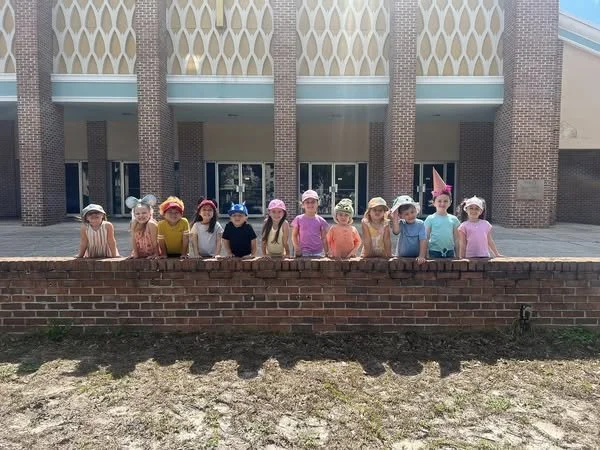A group of twelve young children wearing colorful hats, standing behind a brick wall in front of a building with large windows and brick pillars.