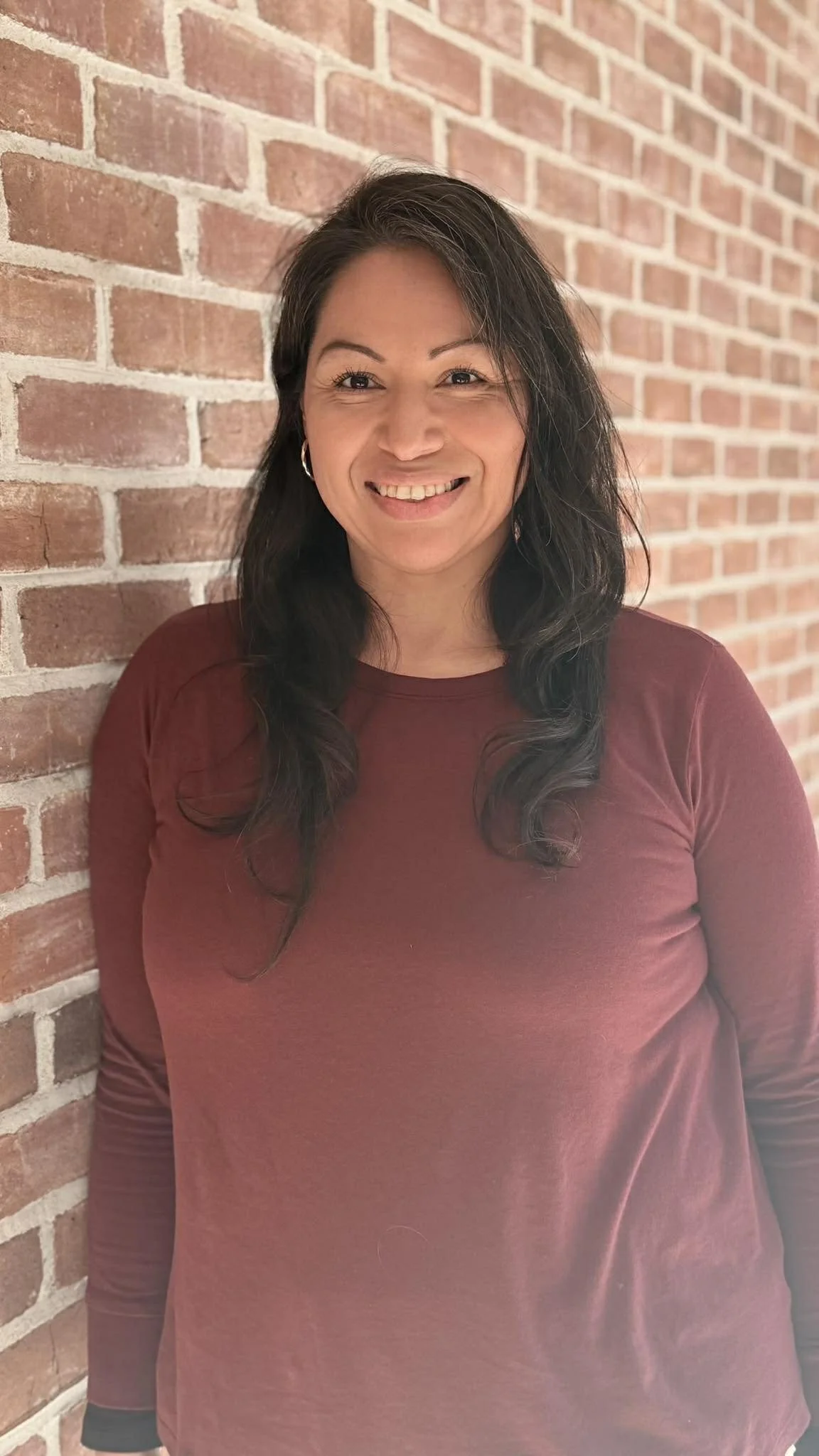 A woman with long dark hair, smiling, standing against a red brick wall, wearing a maroon long-sleeve shirt.