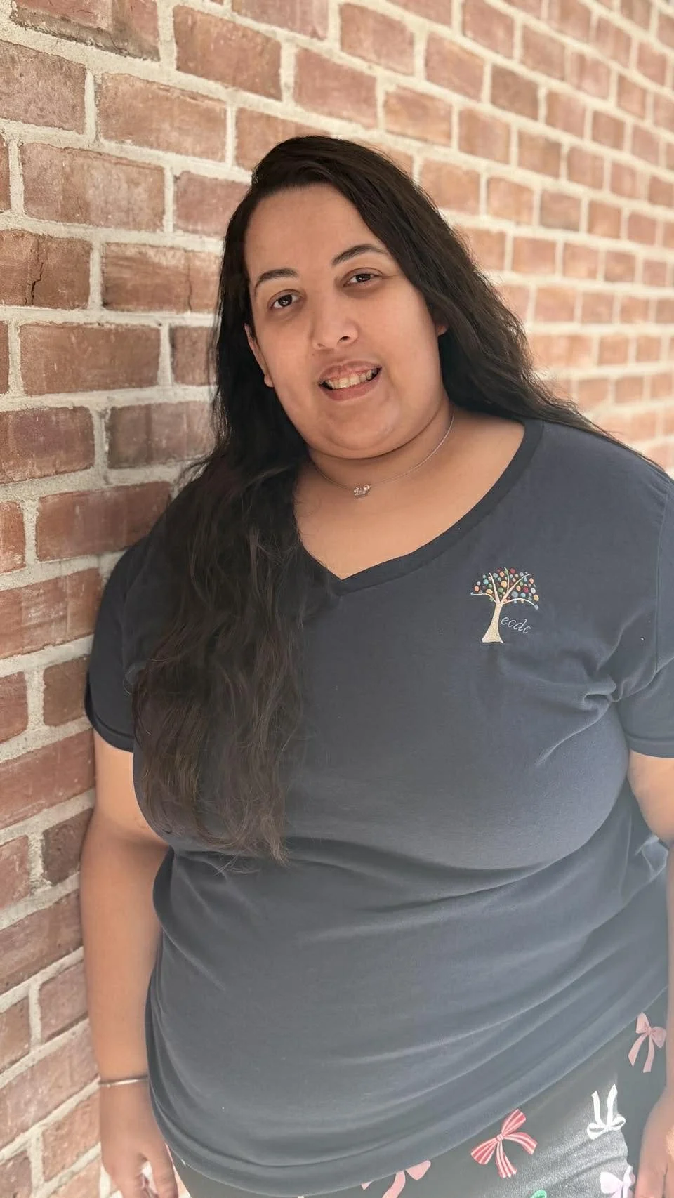 A woman with long dark hair, wearing a dark T-shirt with a colorful tree logo on the left chest, standing against a red brick wall.