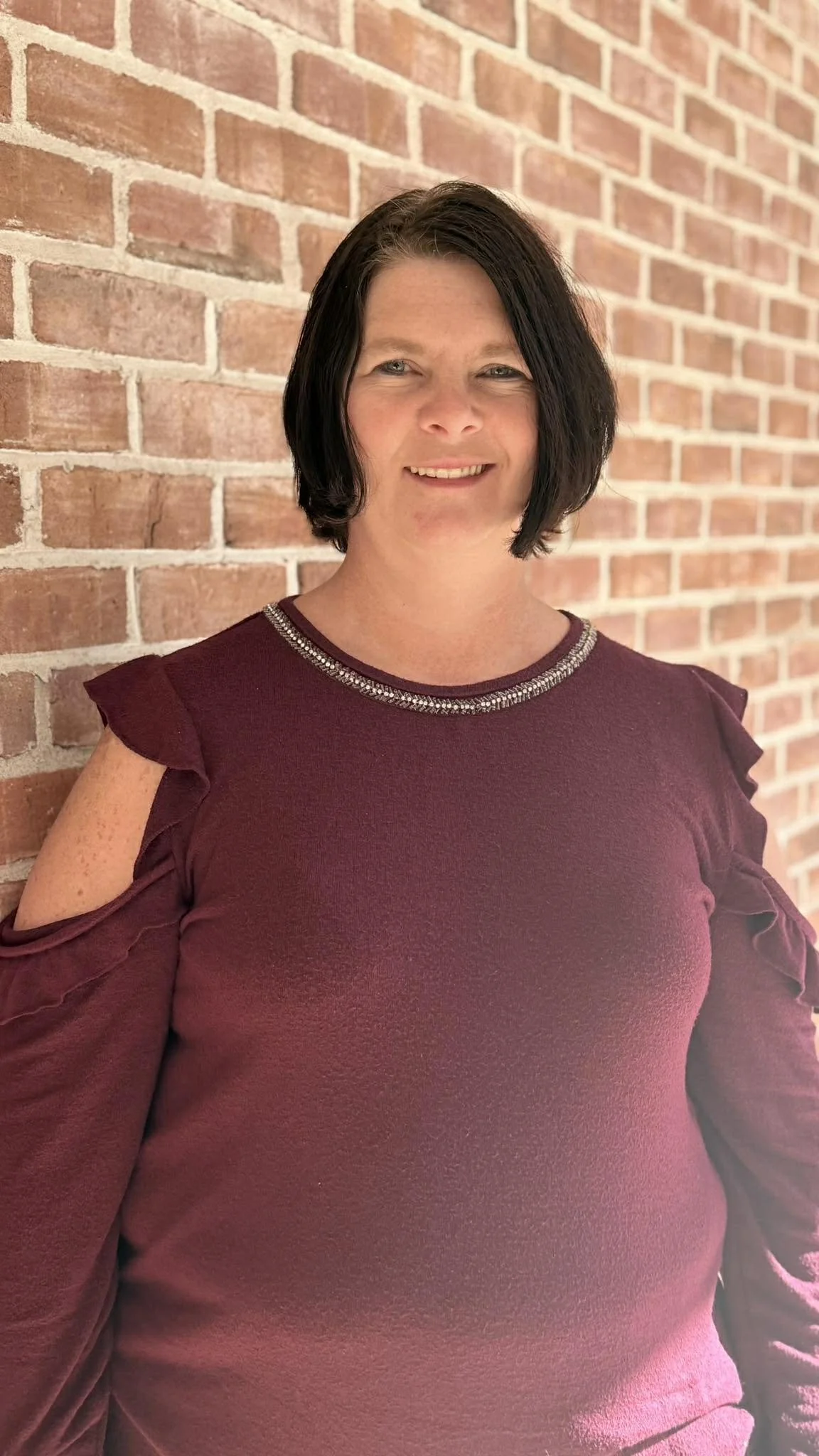 A woman with dark hair smiling and standing against a brick wall, wearing a maroon cold-shoulder top with decorative trim around the neckline.