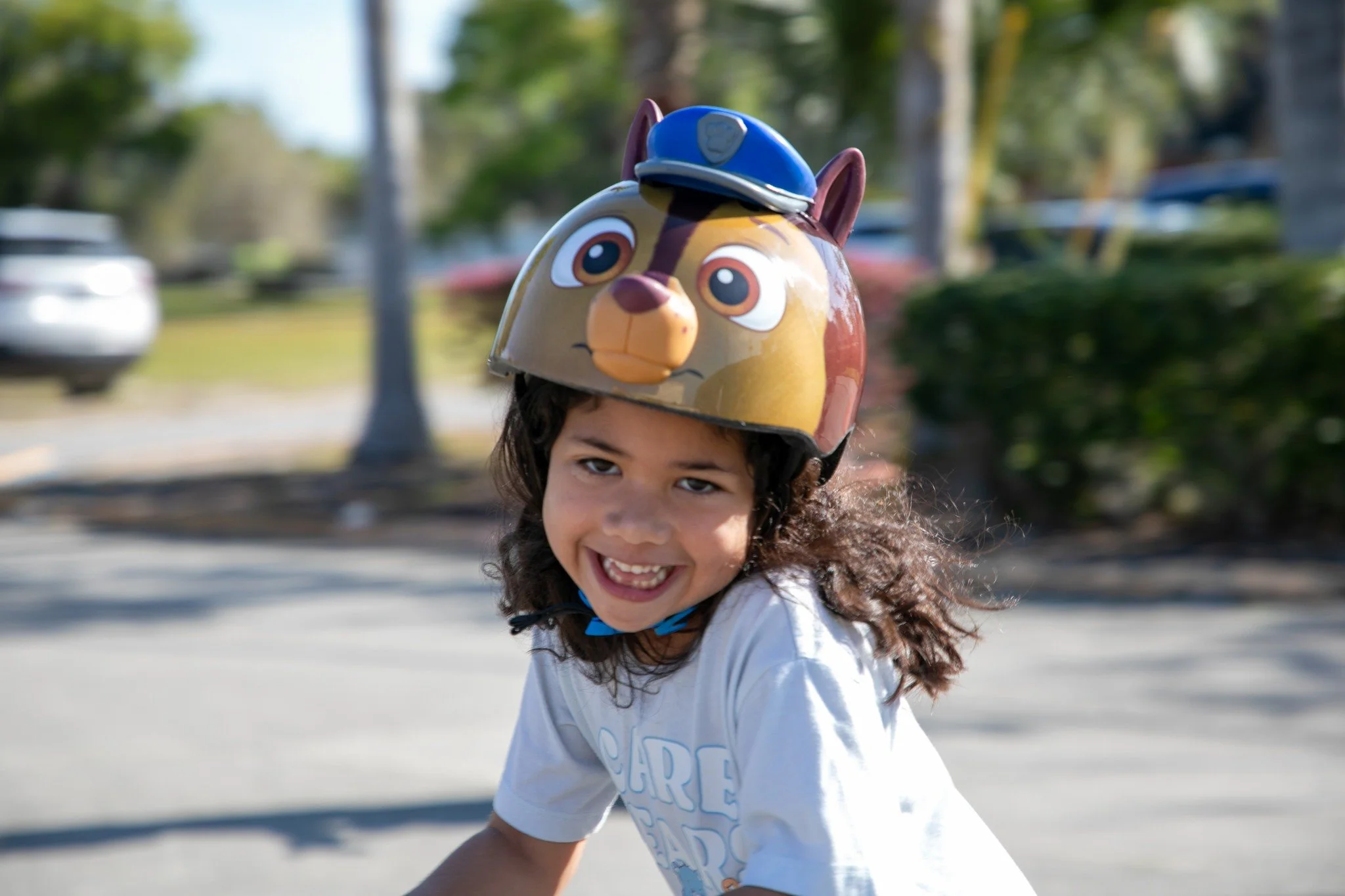 A young girl with curly hair smiling, wearing a white t-shirt, and a helmet resembling Chase from Paw Patrol, outdoors in a park with trees and cars in the background.