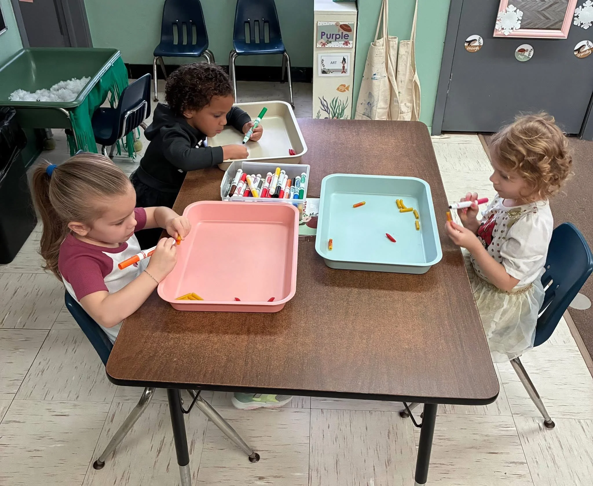 Three young children sitting at a table, coloring with markers. Two girls are on one side, and a girl is on the opposite side. The table has trays with markers. The room has art supplies and backpacks in the background.