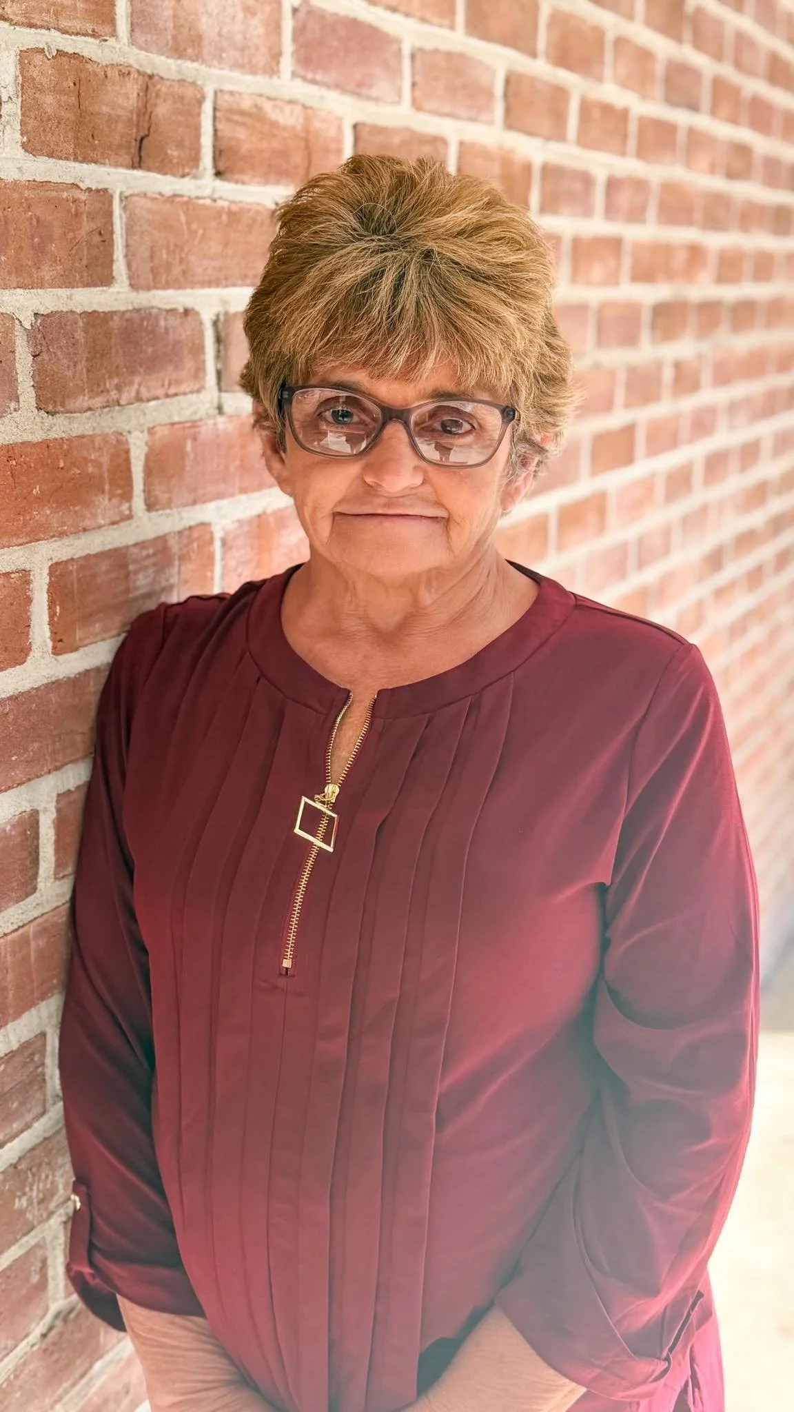 Older woman with short hair, glasses, and a maroon blouse standing near a brick wall.
