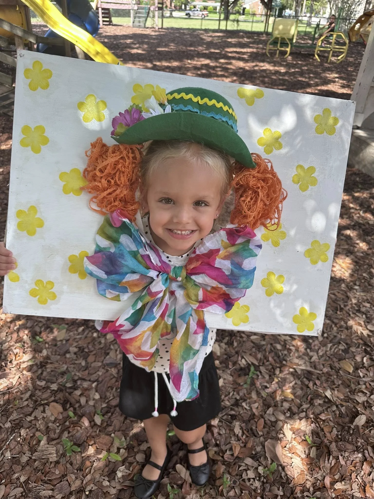 Young girl smiling through a decorated cardboard frame with yellow flower stickers, wearing a colorful bow, a green hat, and black shoes, outdoors on a fallen leaves ground.