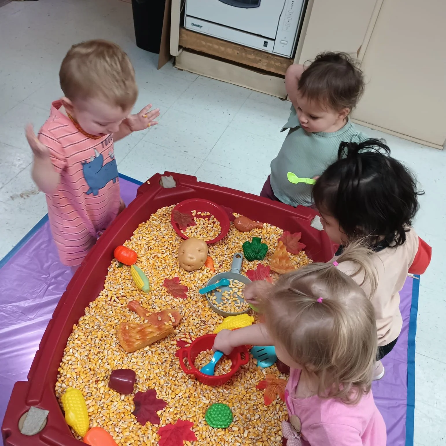 Five children playing in a sensory bin filled with popcorn, plastic vegetables, and toys in a classroom.