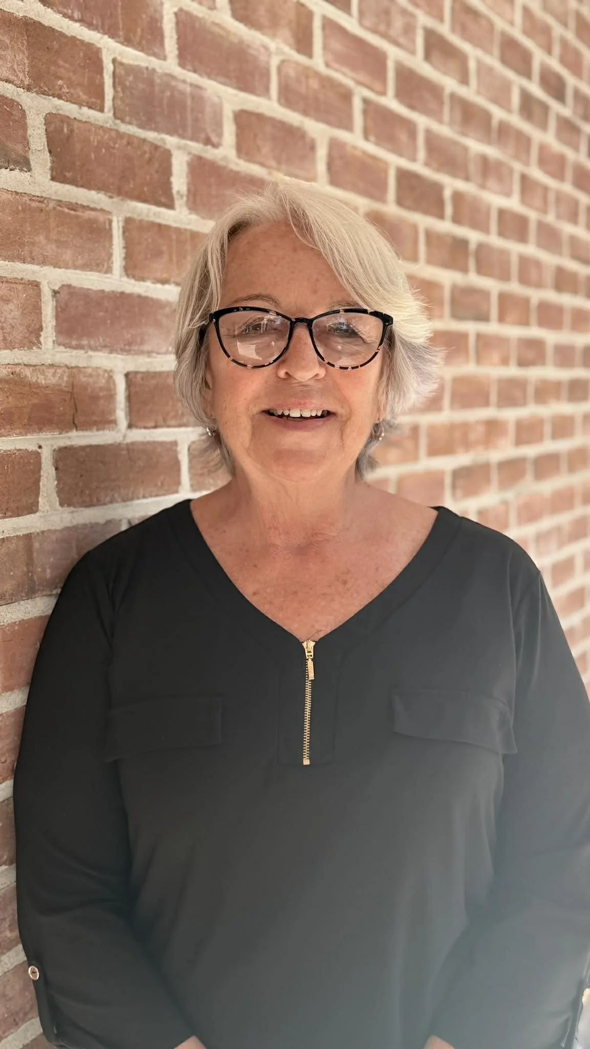 Smiling older woman with short gray hair, wearing glasses and a black top with a gold zipper, standing against a red brick wall.