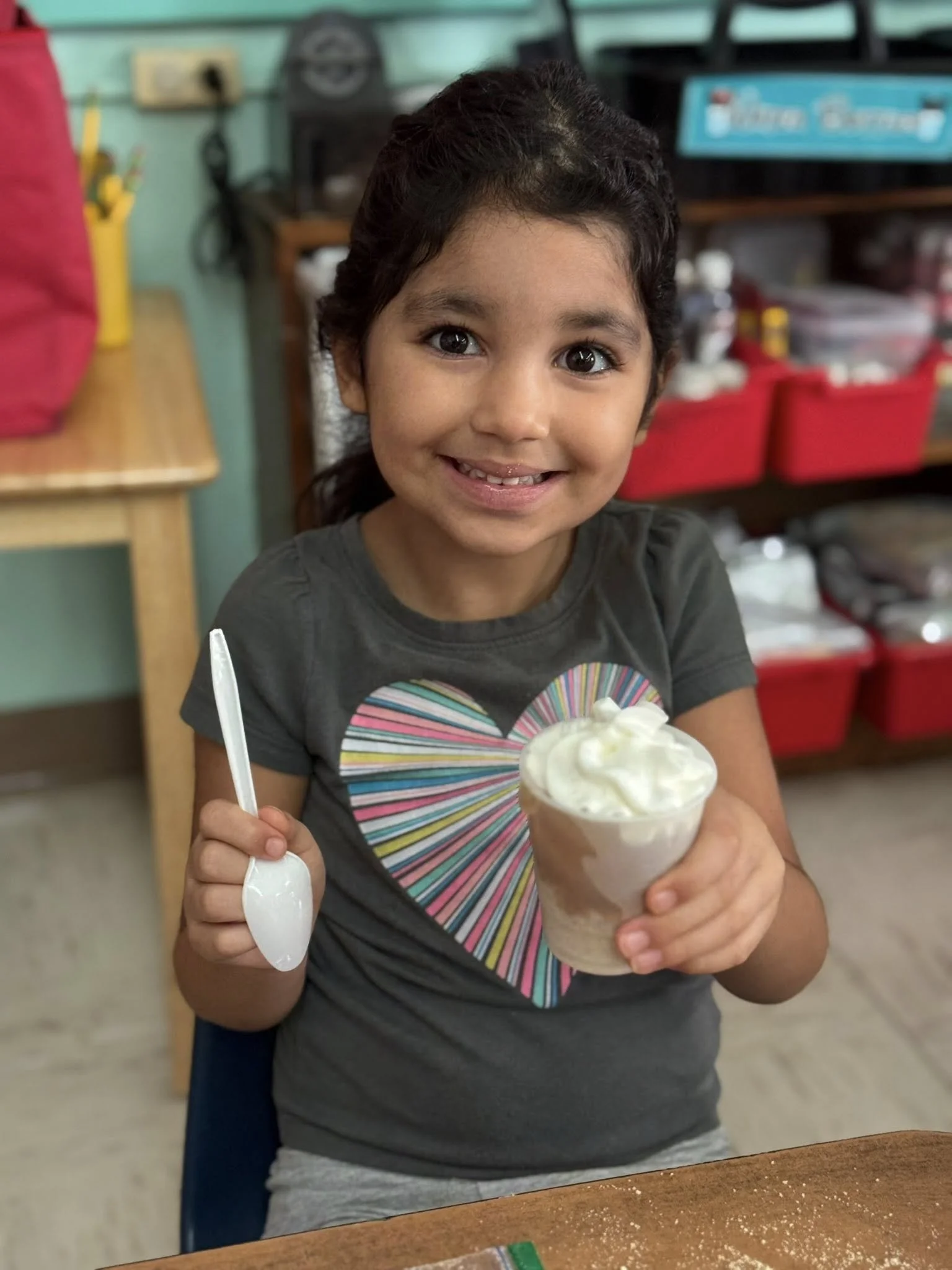 A smiling young girl with dark hair, wearing a gray t-shirt with a colorful heart design, holding a cup of ice cream topped with whipped cream and a plastic spoon inside a cafe or shop.