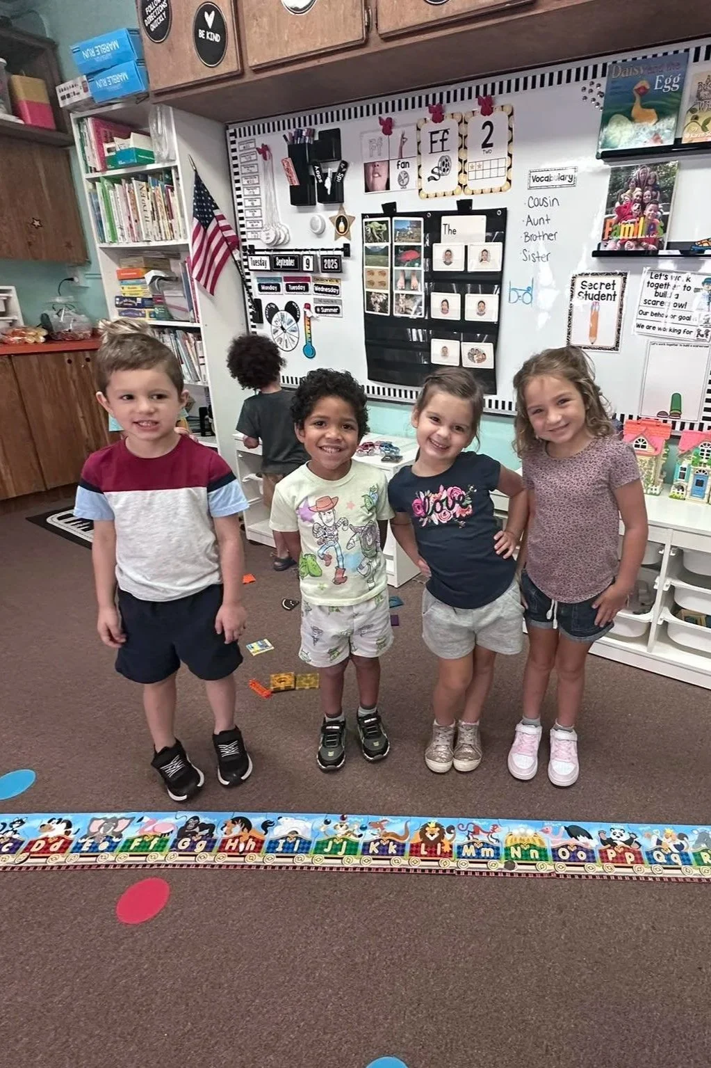 Four preschool children standing together in a classroom, smiling at the camera, with a boy facing away in the background. The classroom has educational posters, a whiteboard, and toys on shelves.