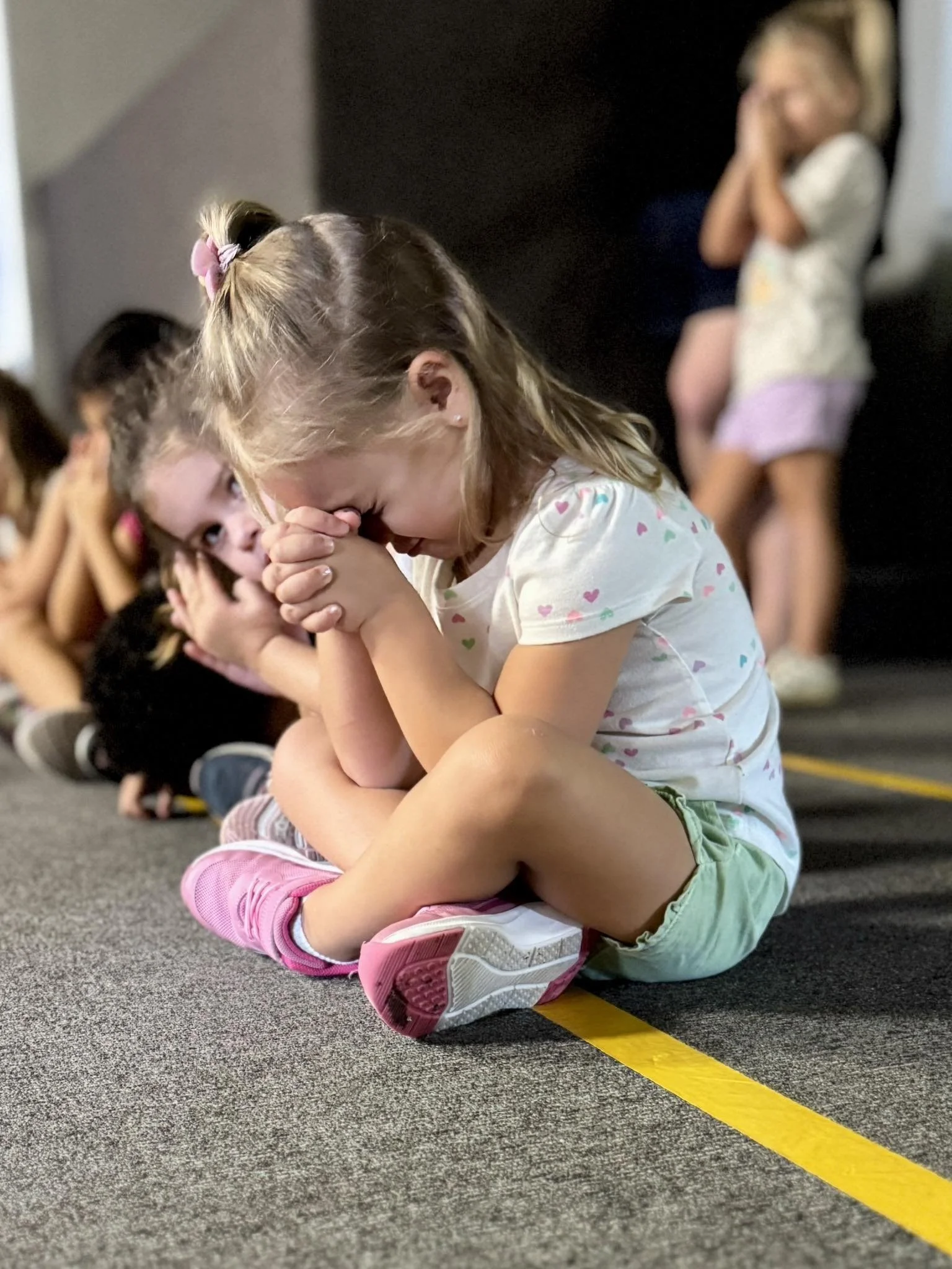 A young girl sitting on a yellow line on a gray carpet, praying or crying with her hands clasped, in a classroom with other children sitting behind her, and another girl in the background covering her ears.