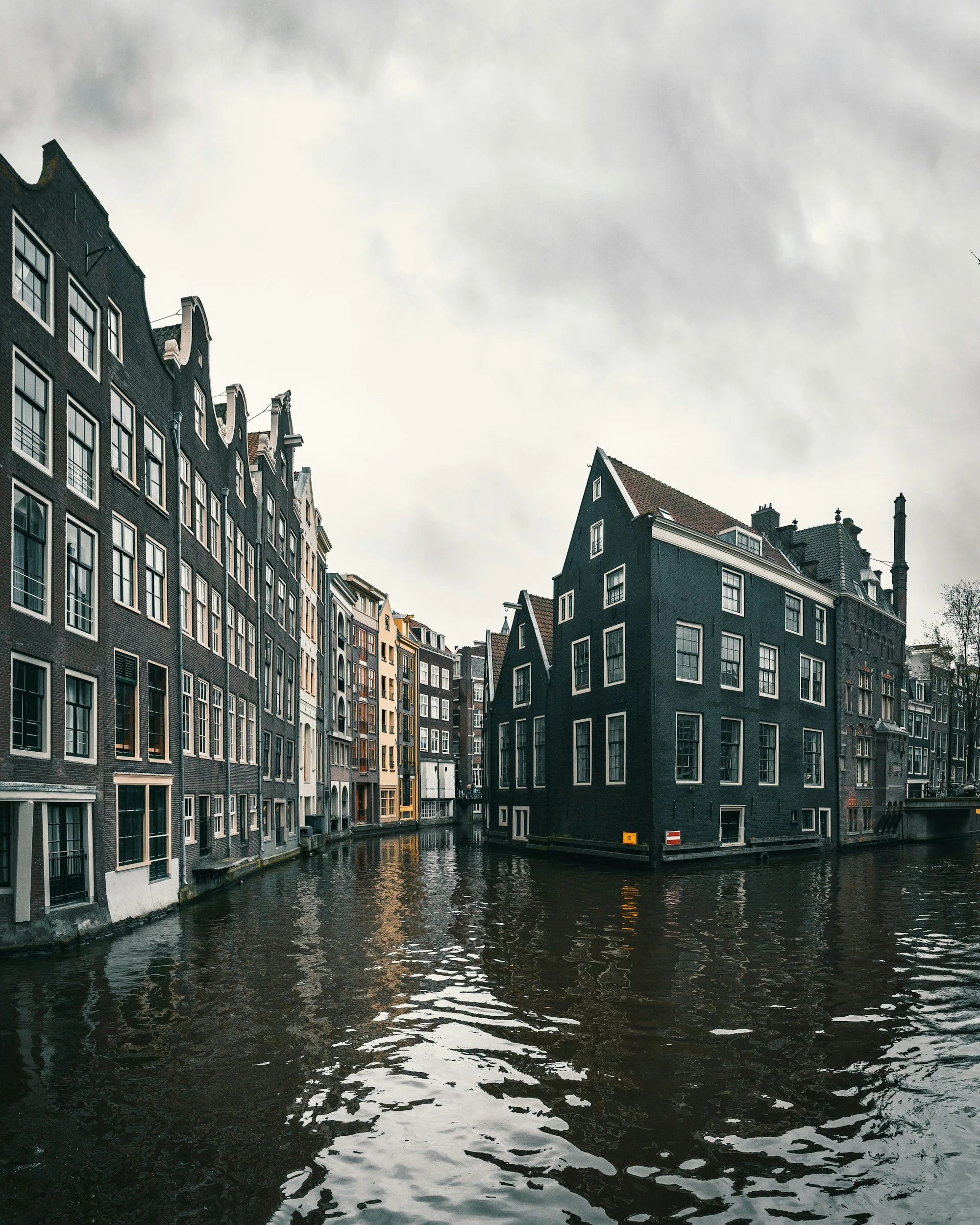 Gothic-style buildings with narrow facades and tall windows situated along a canal under an overcast sky in Amsterdam.