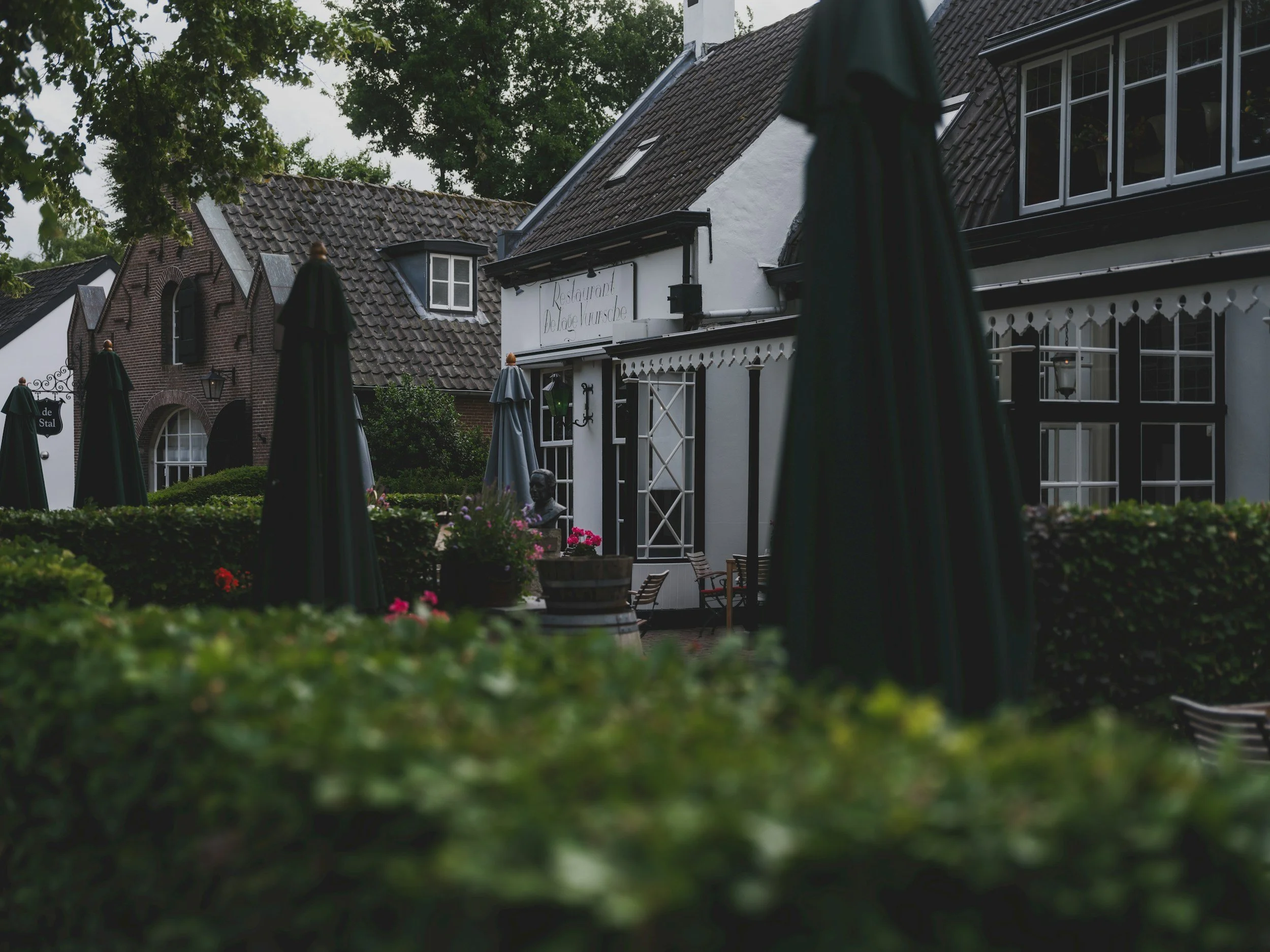 Outdoor patio of a restaurant with closed black umbrellas, potted plants, garden statues, and a white building with decorative trim and large windows.