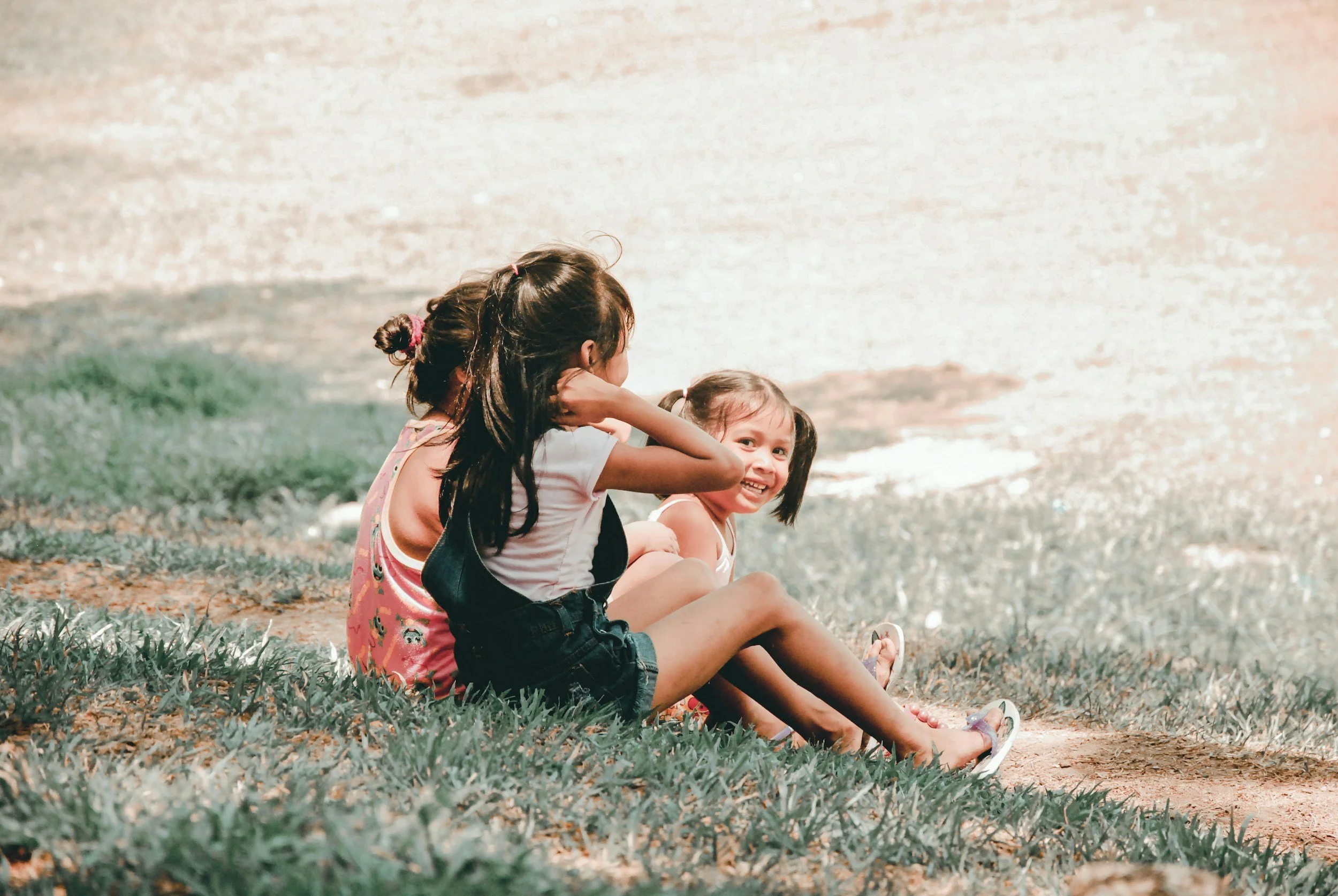 Three young girls sitting on the grass and talking outdoors, with a blurred background and warm sunlight.