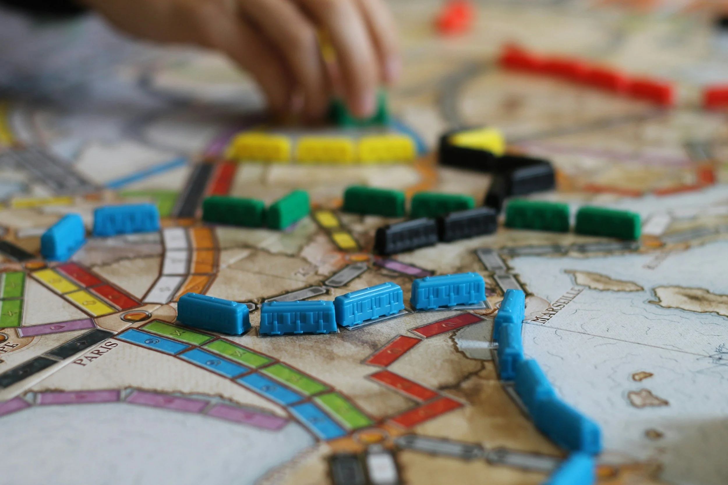 Close-up of a Monopoly game board with colorful plastic player pieces, property tiles, and game tokens, with a person's hand moving a game piece in the background.