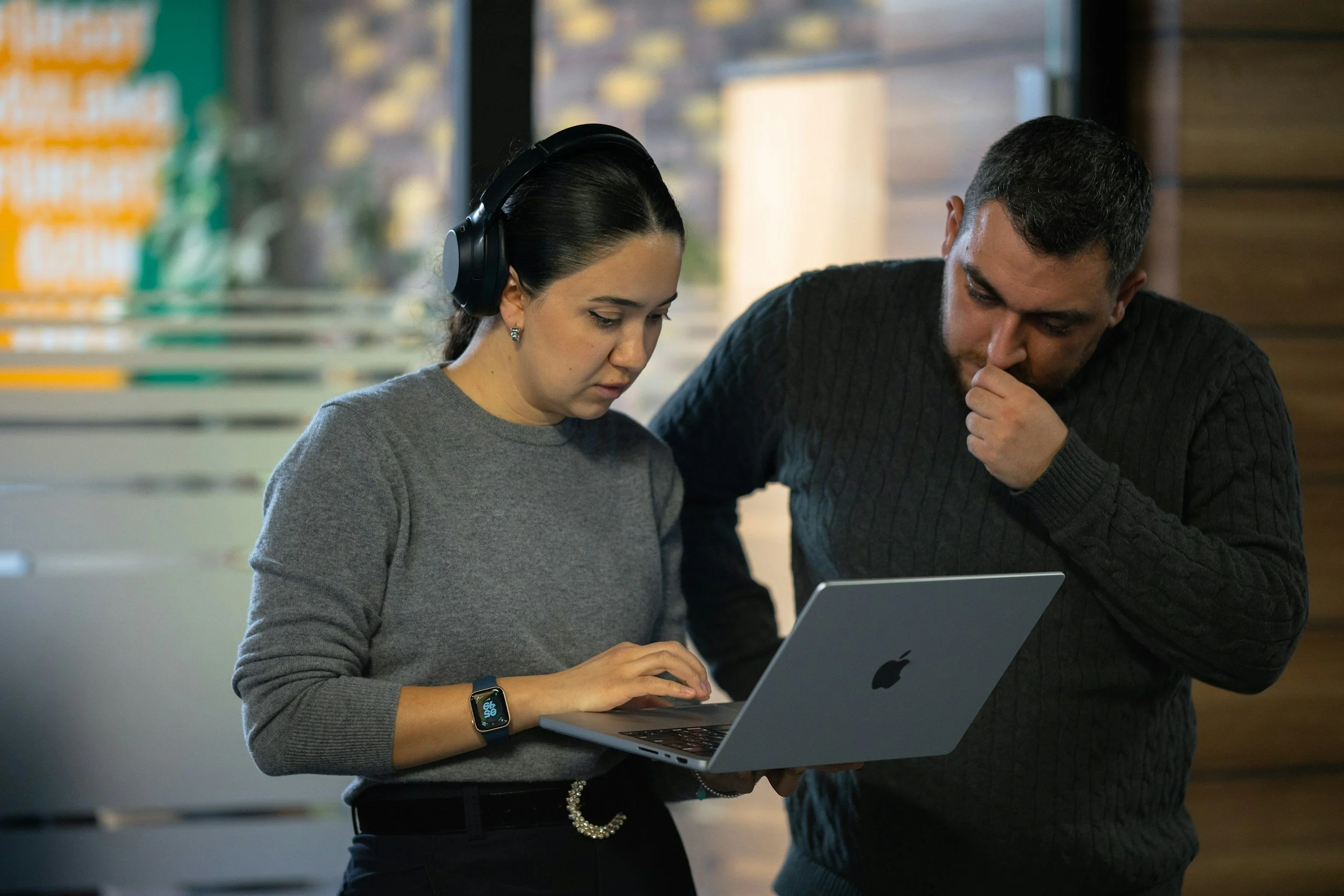 A woman wearing headphones and a smartwatch looks at a laptop screen while a man in a dark sweater leans over to look at it, both appearing focused.