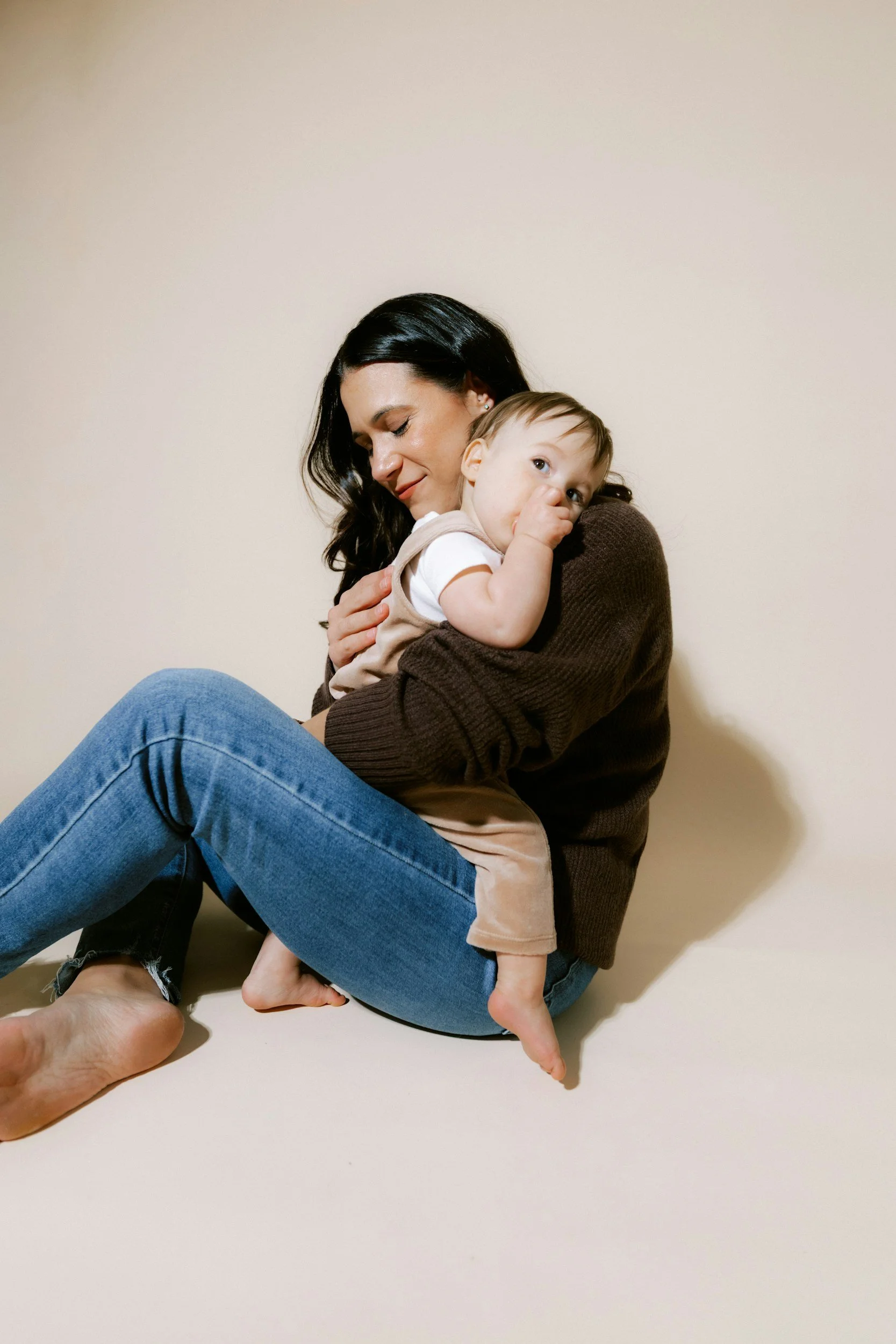 A woman with dark hair holding a young child with light hair, both sitting on the floor against a plain beige wall. The woman appears to be smiling softly with eyes closed, while the child is looking at the camera with a thumb in their mouth.