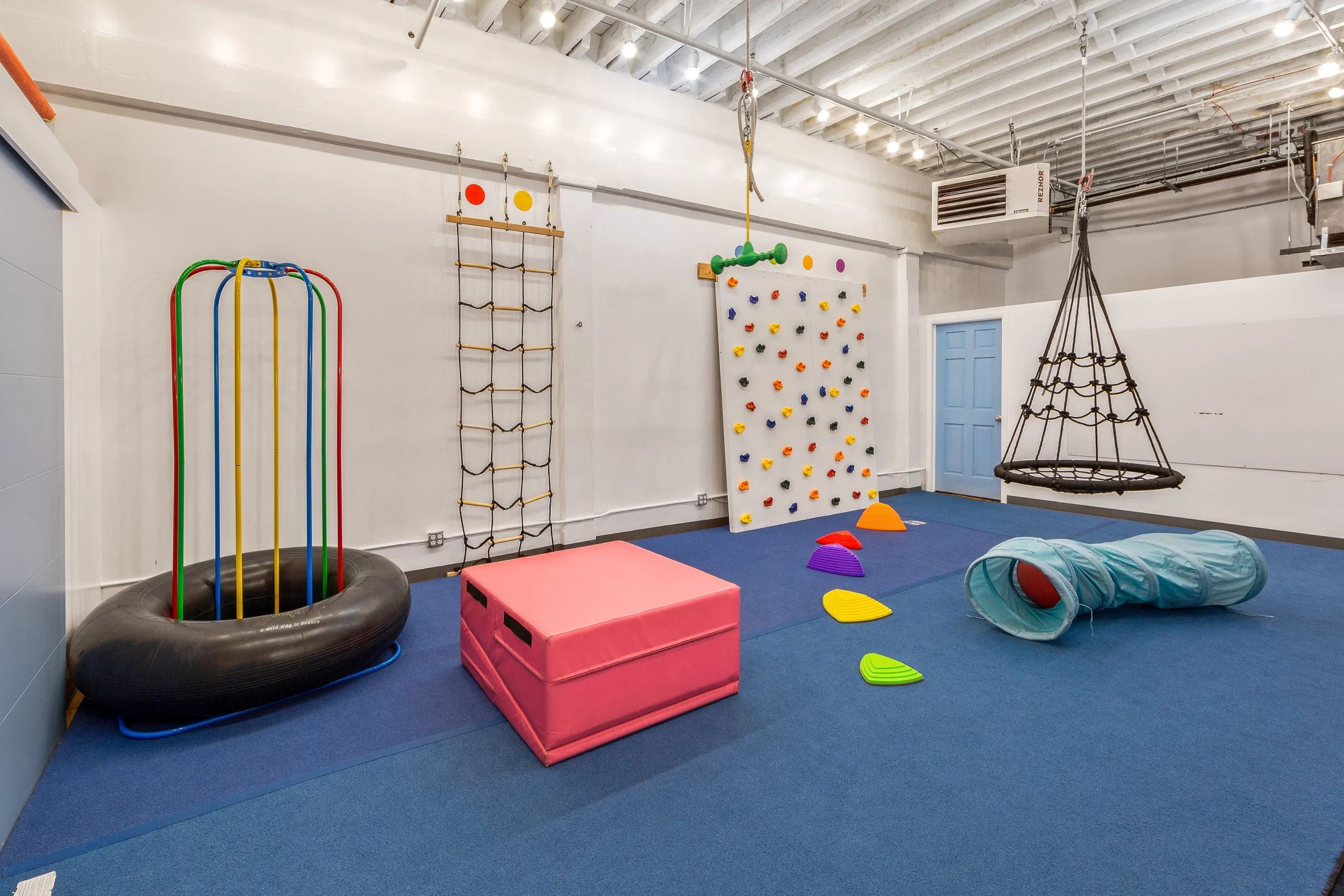 Indoor play area with climbing wall, swings, soft blocks, tunnel, and colorful stepping stones on blue carpet.