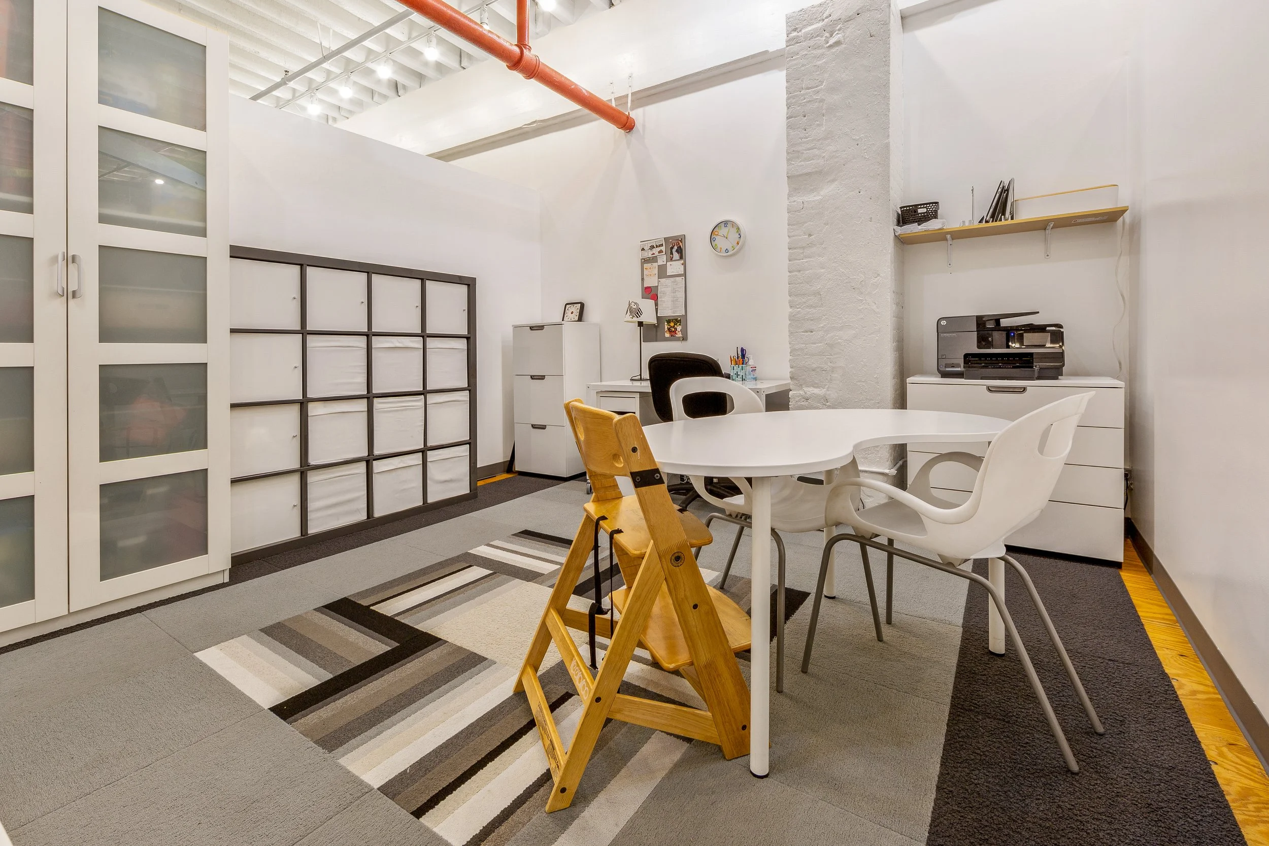 An office space with white walls, a rectangular white table, and various chairs including white plastic and a wooden high chair. There are cabinets, a filing cabinet, a printer, a shelf with books, and a bulletin board on the wall. The ceiling has exposed beams and an orange pipe, and the floor is covered with gray, beige, black, white, and striped carpets.