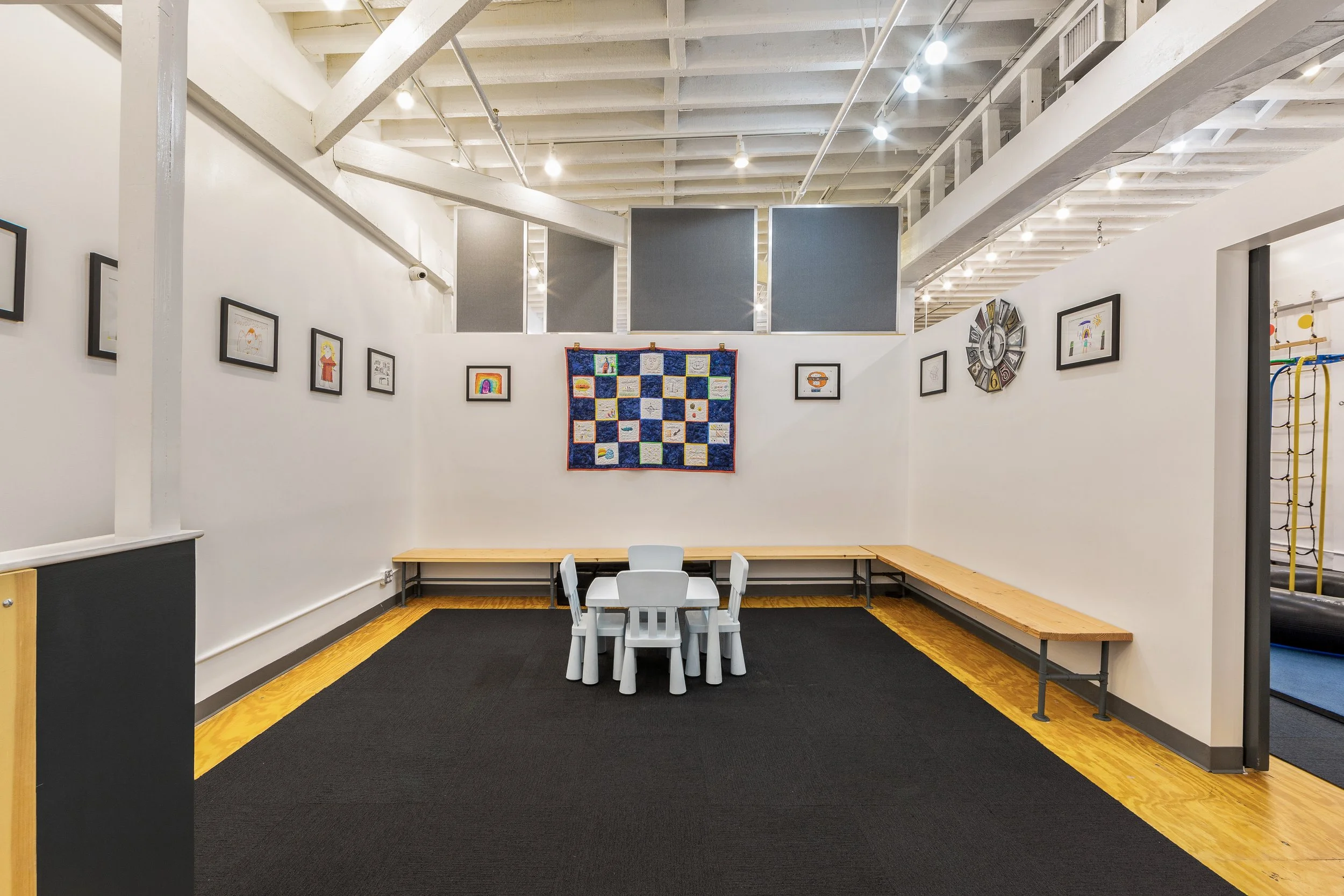An art display in a gallery with framed artwork on white walls, a quilt in the center, a small table with stools, and a bench around the room.