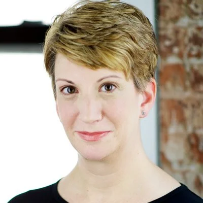 A woman with short, light brown hair, wearing a black top, smiling slightly, with a brick wall and window in the background.