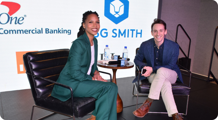 Two people sitting on stage in front of a large screen, smiling, during a panel discussion or interview. The woman on the left wears a green suit, and the man on the right wears a blue blazer, light-colored pants, and brown shoes. There are water bottles and microphones on a small table between them.