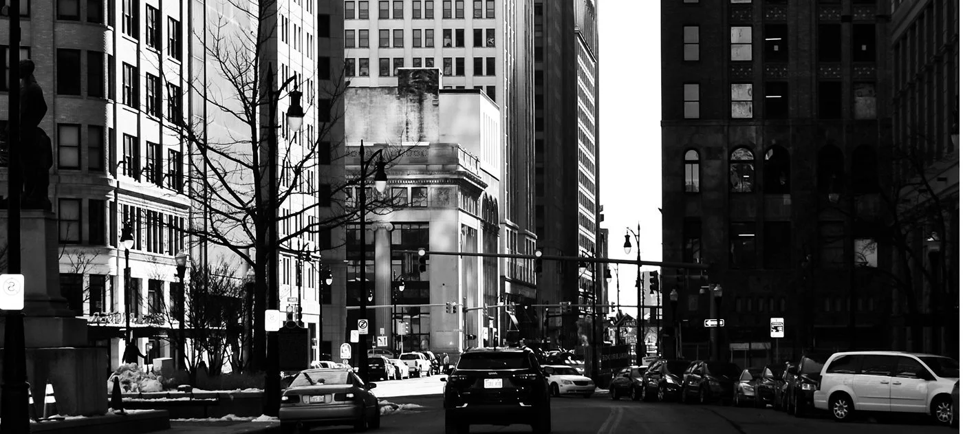 Black and white photo of city street with parked cars, leafless trees, and tall buildings on both sides, with a bright sky in the background.