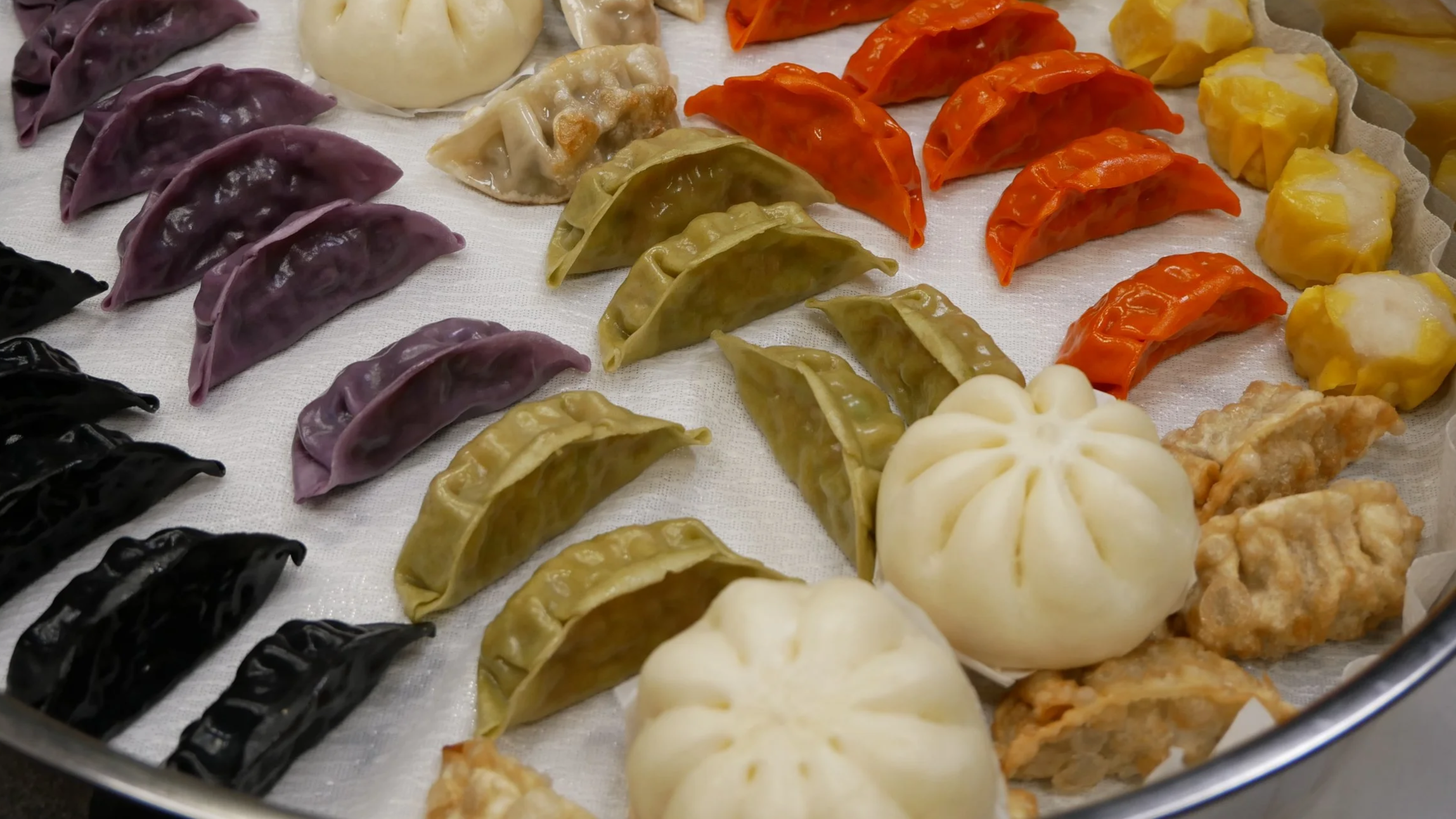 Colorful assortment of steamed dumplings wrapped in various colored doughs including black, purple, green, orange, yellow, and white, arranged on a white cloth-lined tray.