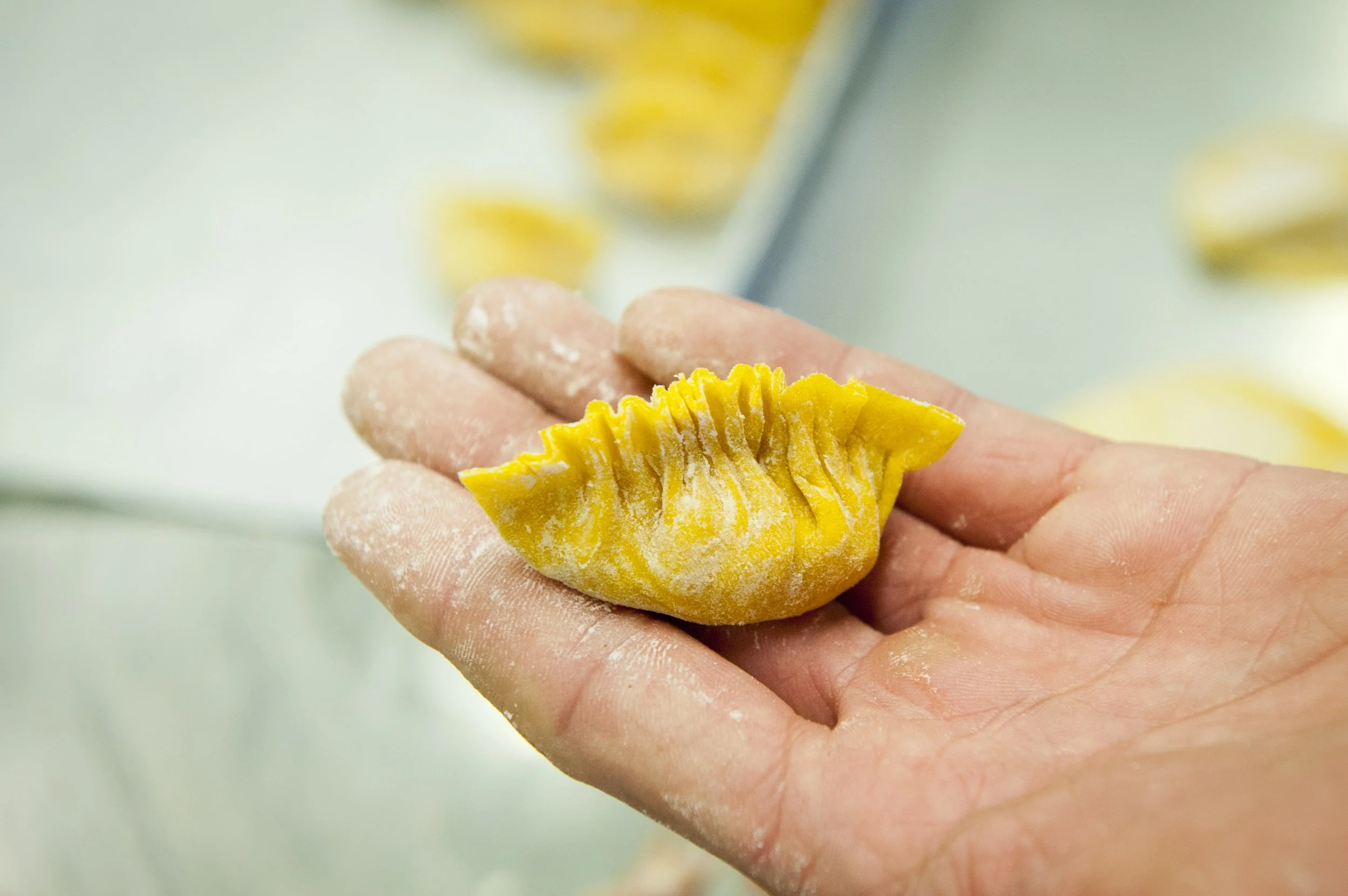 A hand holding a partially made yellow dumpling with crimped edges, with a tray of more dumplings in the background.