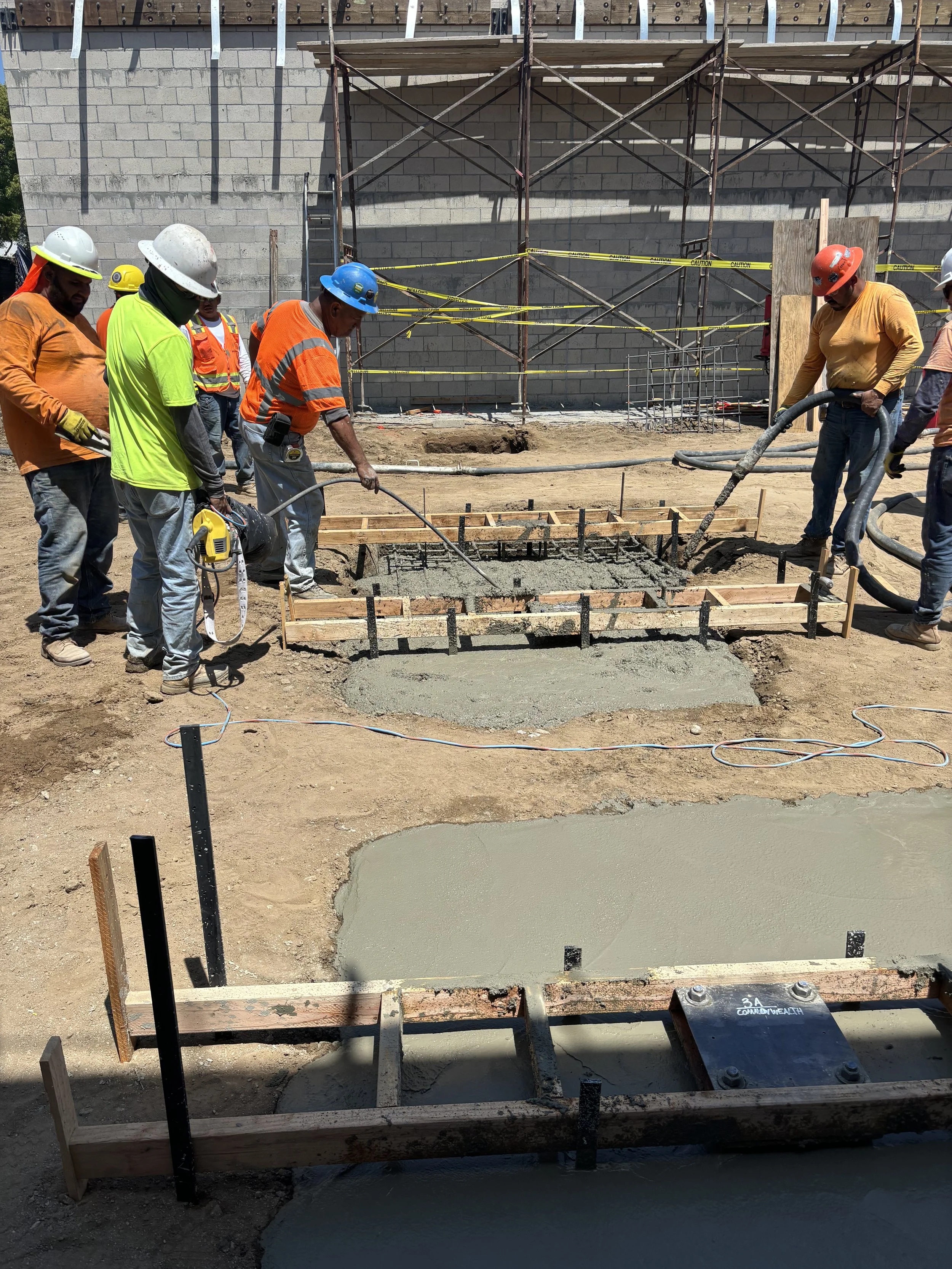 Construction workers pouring and finishing concrete on a building site, with scaffolding and a cinder block wall in the background.