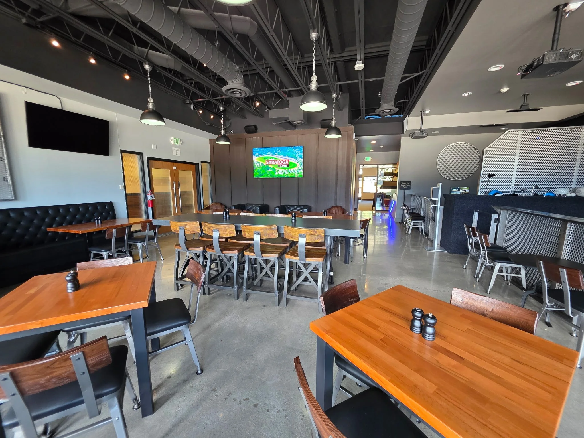 Interior of a restaurant or bar with wooden tables, chairs, a TV screen showing 'Saratoga', and a black and gray decor with modern lighting.