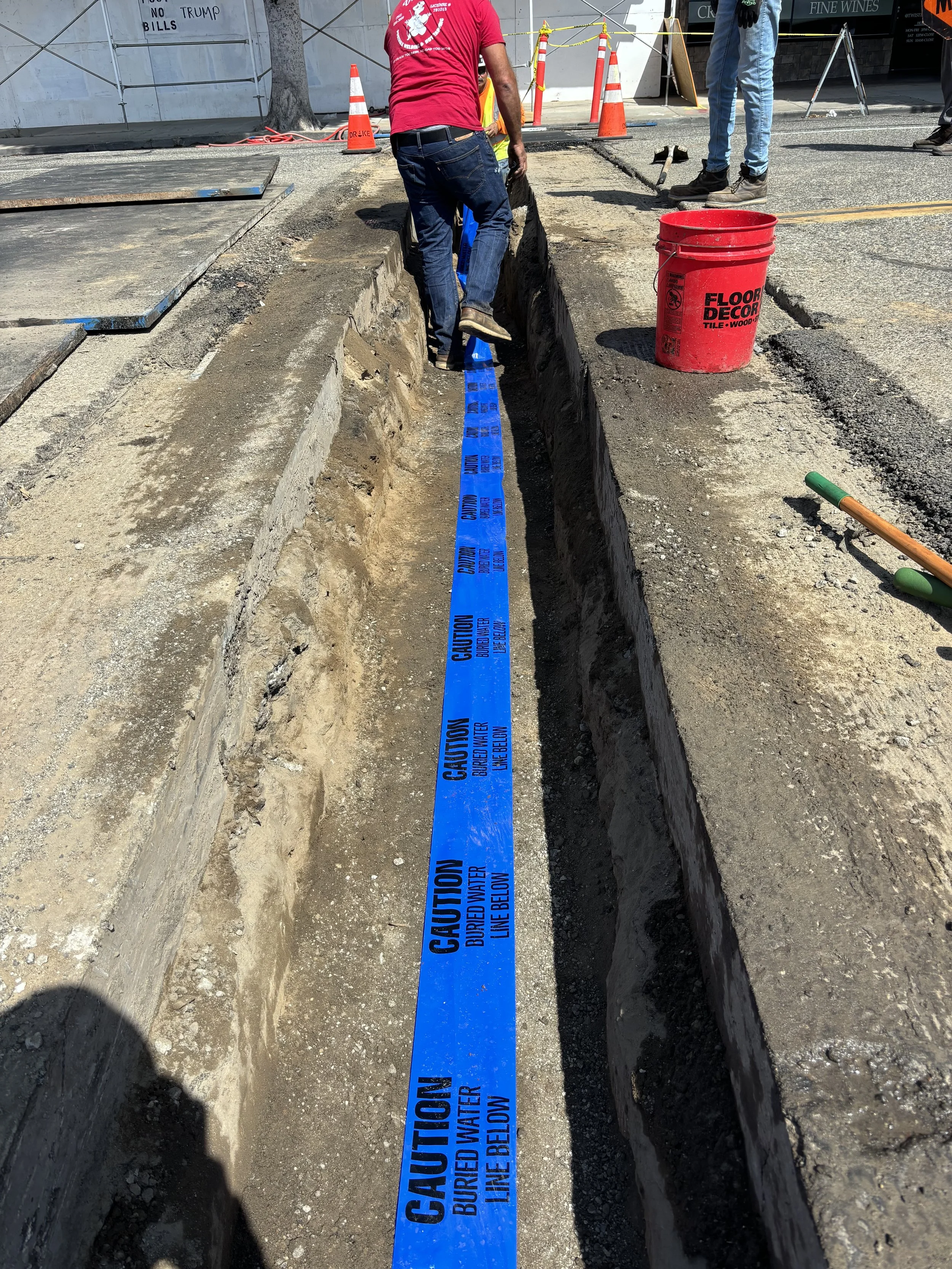 Construction workers laying down blue caution tape in a trench on a city street, surrounded by orange cones and construction equipment.