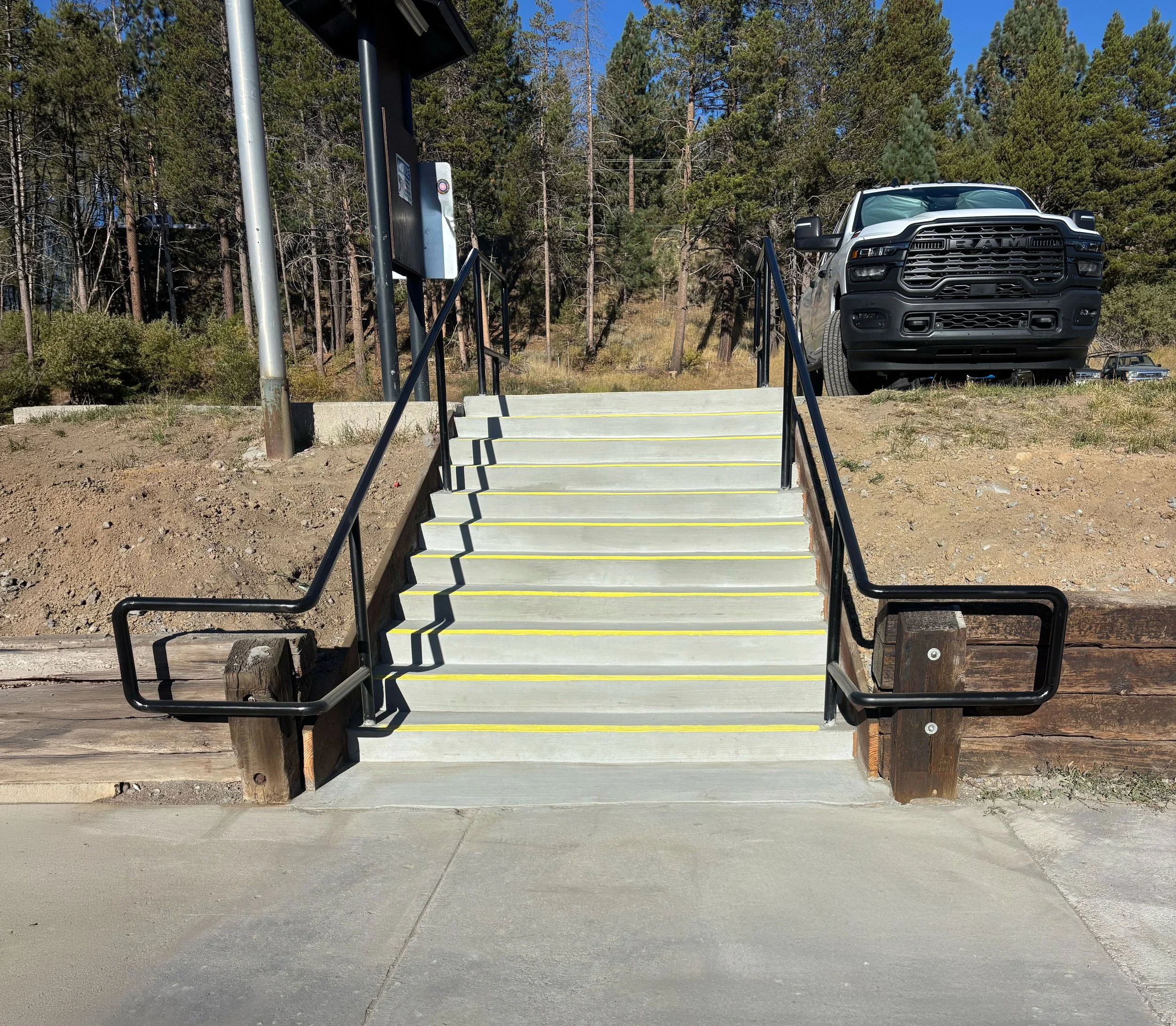 Concrete stairs with black railings and yellow safety lines leading up to a parking lot, on a sunny day with trees in the background and a parked black truck at the top.