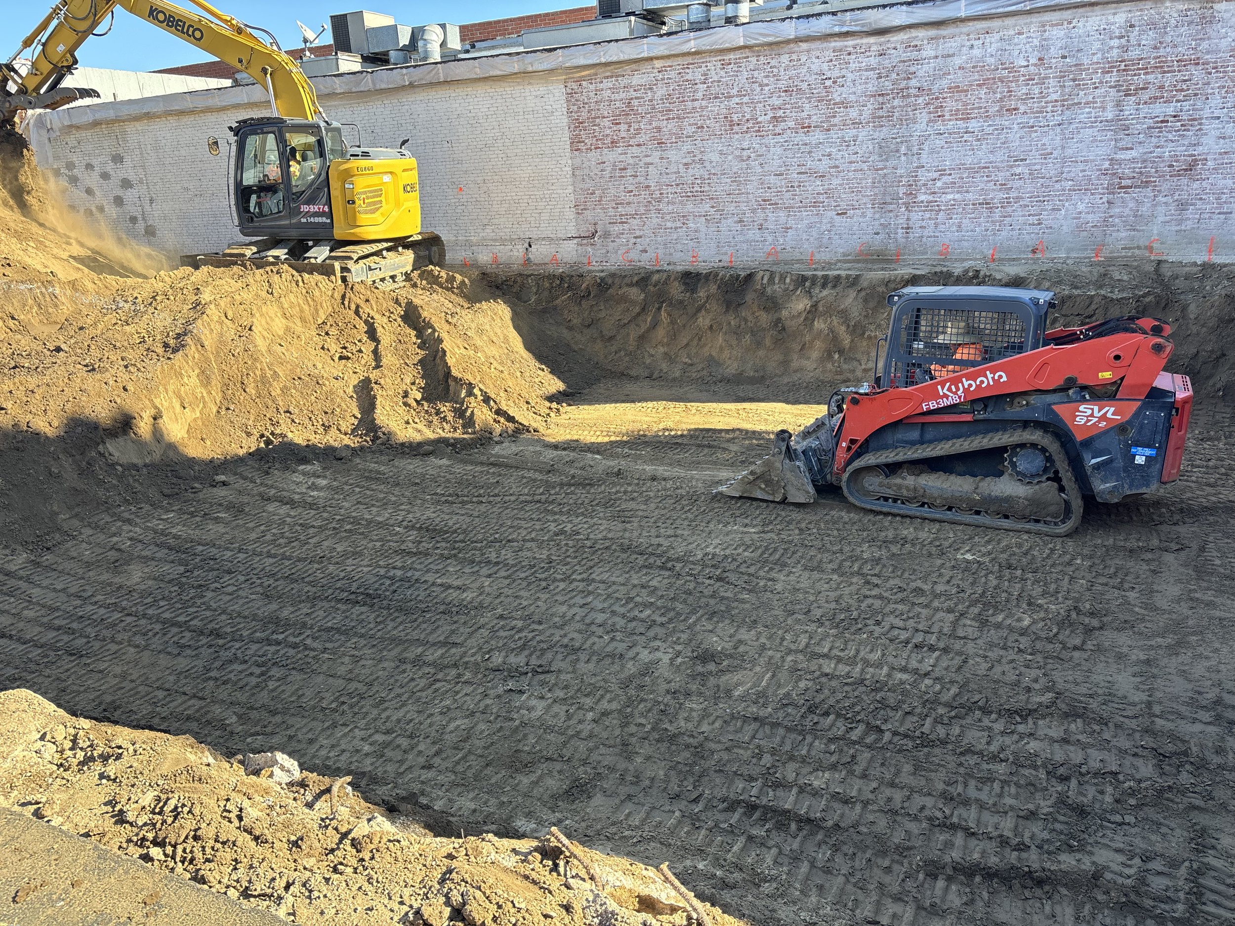 Construction site with a small red Kubota skid-steer loader and a yellow Komatsu excavator working on dirt excavation next to a brick wall.