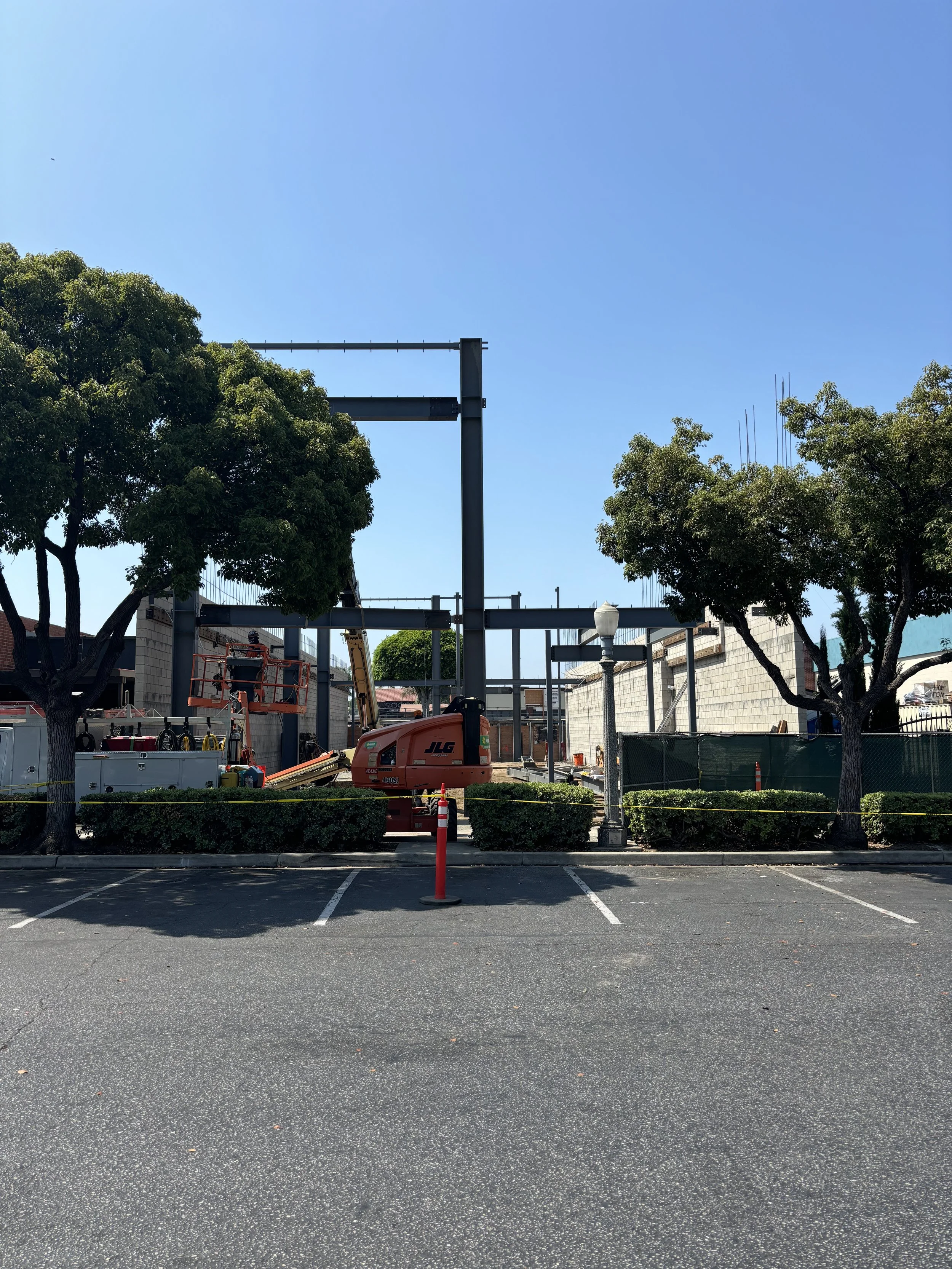 Construction site with metal framework, trees, and construction equipment, view from parking lot.