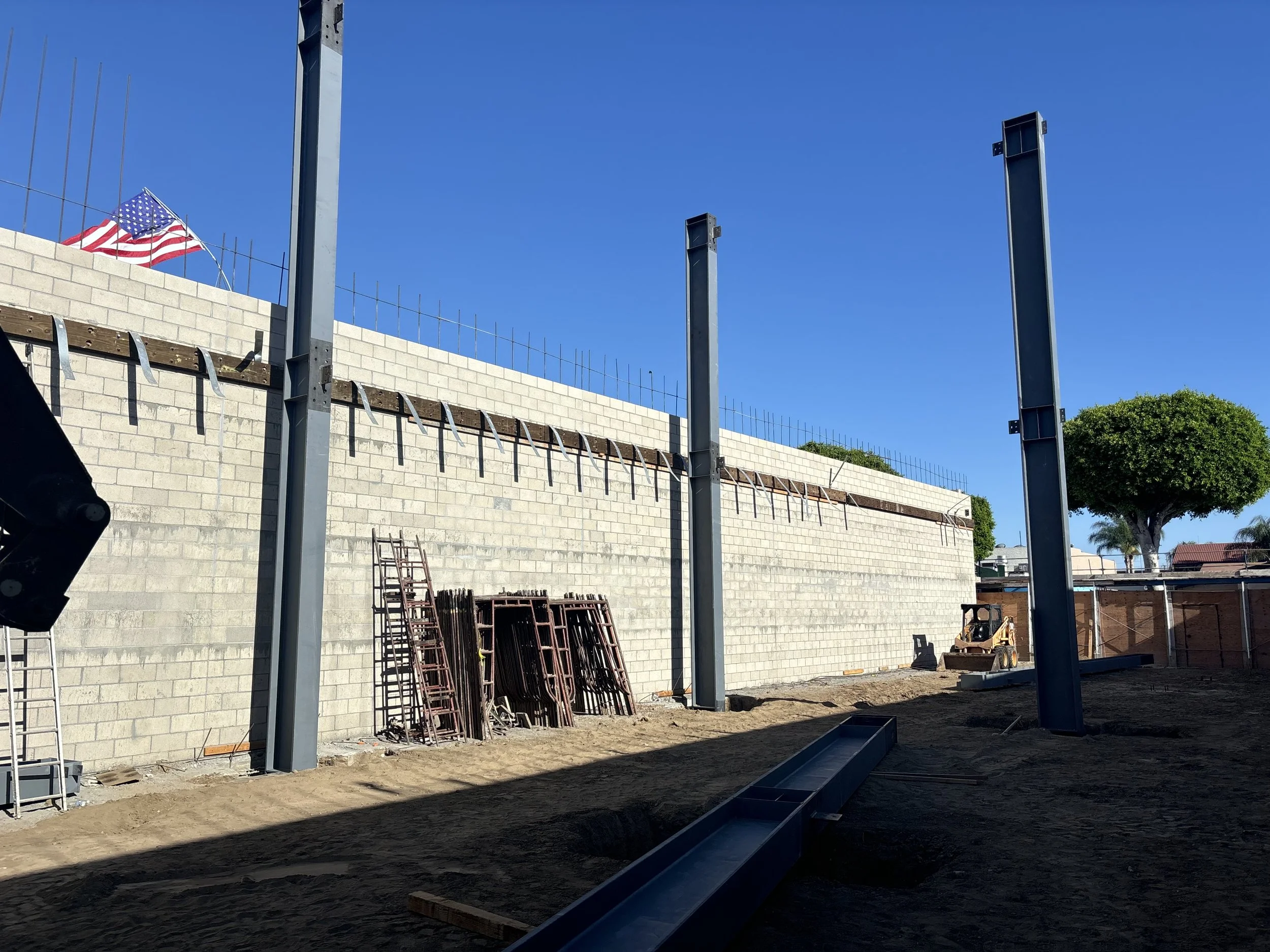 Construction site with steel support beams, a concrete block wall, a ladder, construction materials, and an American flag on the top left corner under a clear blue sky.